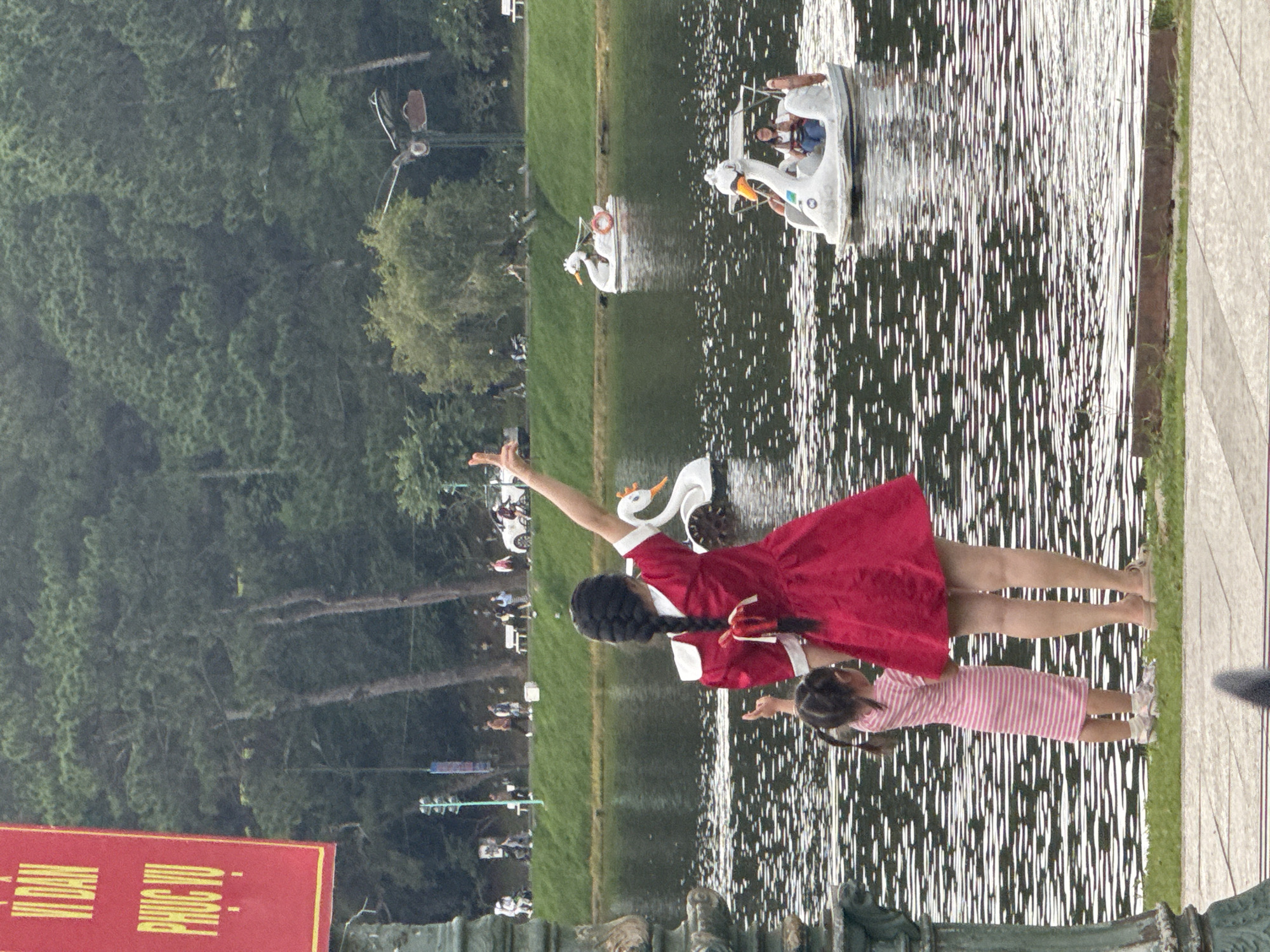 The image captures a serene scene at Xuan Huong Lake in Da Lat, Vietnam. In the foreground, a woman in a red dress and a young girl in a pink striped dress stand on the grassy bank, looking out at the water. The woman has her arm raised, and the girl appears to be pointing at something in the lake.
The lake is dotted with several swan-shaped pedal boats. One boat in the mid-ground contains two individuals, while another further back is empty. The background features a lush green landscape with tall trees, suggesting the surrounding parkland. A red banner with Vietnamese text is visible on the left side of the frame, and other park-goers can be seen in the distance walking along a paved path. The water's surface is rippled, reflecting the overcast sky and the surrounding greenery.