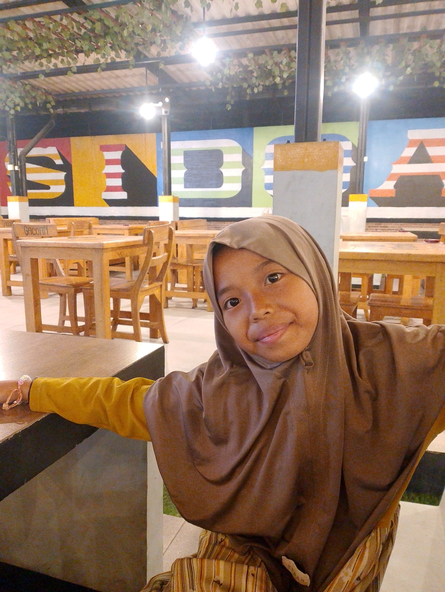 In Bringinbendo, Indonesia, a young girl wearing a brown hijab and a mustard-yellow long-sleeved shirt poses for a photo. She is seated at a wooden table in an outdoor or semi-outdoor dining area. Behind her, a colorful mural adorns the wall, featuring abstract shapes and stripes in bold colors like red, yellow, blue, and green. Wooden tables and chairs are neatly arranged throughout the space, suggesting a restaurant or cafe setting. The ceiling is covered with foliage, and artificial lighting illuminates the area. The girl smiles warmly at the camera, her right arm extended, resting on the edge of the table. A bracelet with pink beads is visible on her wrist. The overall atmosphere is casual and inviting.