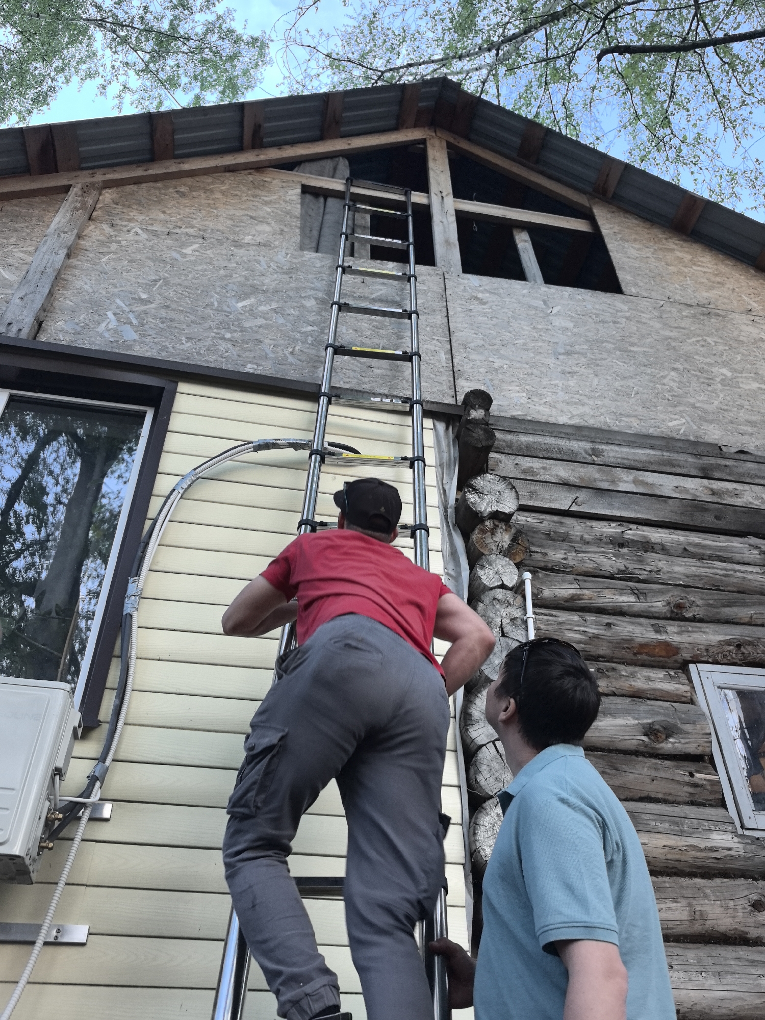 The image depicts two men working on the exterior of a two-story building in what is reported to be Noviy, Russia. The building has a distinct architectural style, combining elements of both traditional log cabin construction and modern siding and roofing materials. The lower portion of the structure is constructed from roughly hewn logs, typical of log cabins, while the upper portion is covered with what appears to be oriented strand board (OSB). The roof is made of corrugated metal.
A man wearing a red shirt, gray pants, and a dark baseball cap is ascending an extension ladder leaning against the building. He is several rungs up, his back to the camera. Below him, another man in a blue shirt is looking up at the man on the ladder.
The ladder is positioned near the juncture of the log cabin and siding sections of the building, next to an air conditioning unit mounted on the siding. A protective conduit or tubing runs alongside the siding, partially obscured by the ladder. The building also includes framed windows on both the log cabin and siding sections. Tree branches and sky are visible above the building.