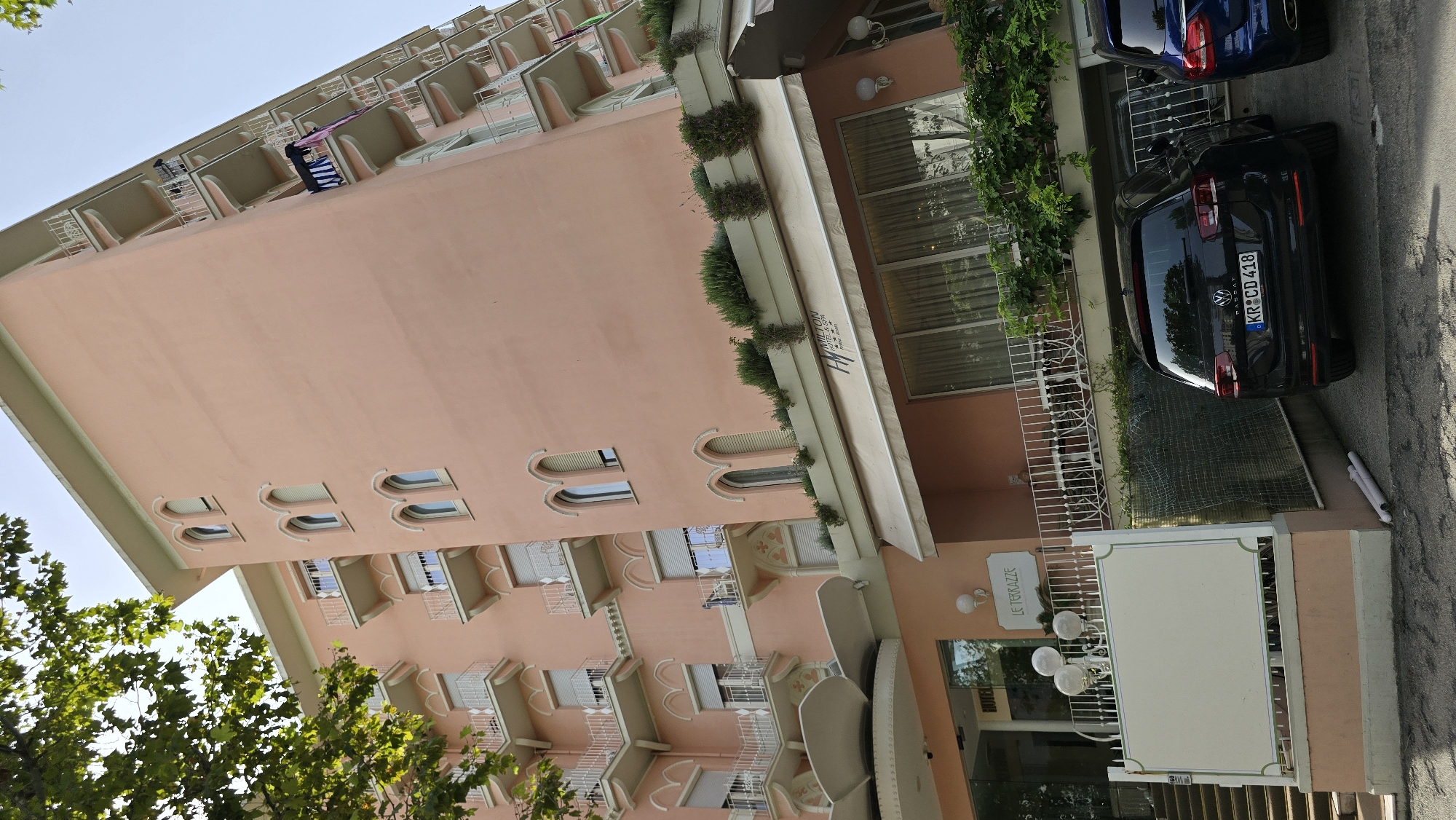The image shows the facade of the Hamilton Hotel & Spa in Rimini, Italy. The hotel is a tall, peach-colored building with multiple balconies and arched windows. A black Volkswagen Passat with a German license plate is parked in front of the hotel, along with a blue SUV. The hotel has a sign that reads "LE TERRAZZE" and another sign with the hotel's name and logo. Trees are visible in the upper left corner of the image, and the sky is clear and blue. The entrance to the hotel is visible on the left side of the image, with a set of stairs leading up to it. The hotel appears to be a well-maintained establishment, and the overall scene is one of a pleasant day in an Italian city.