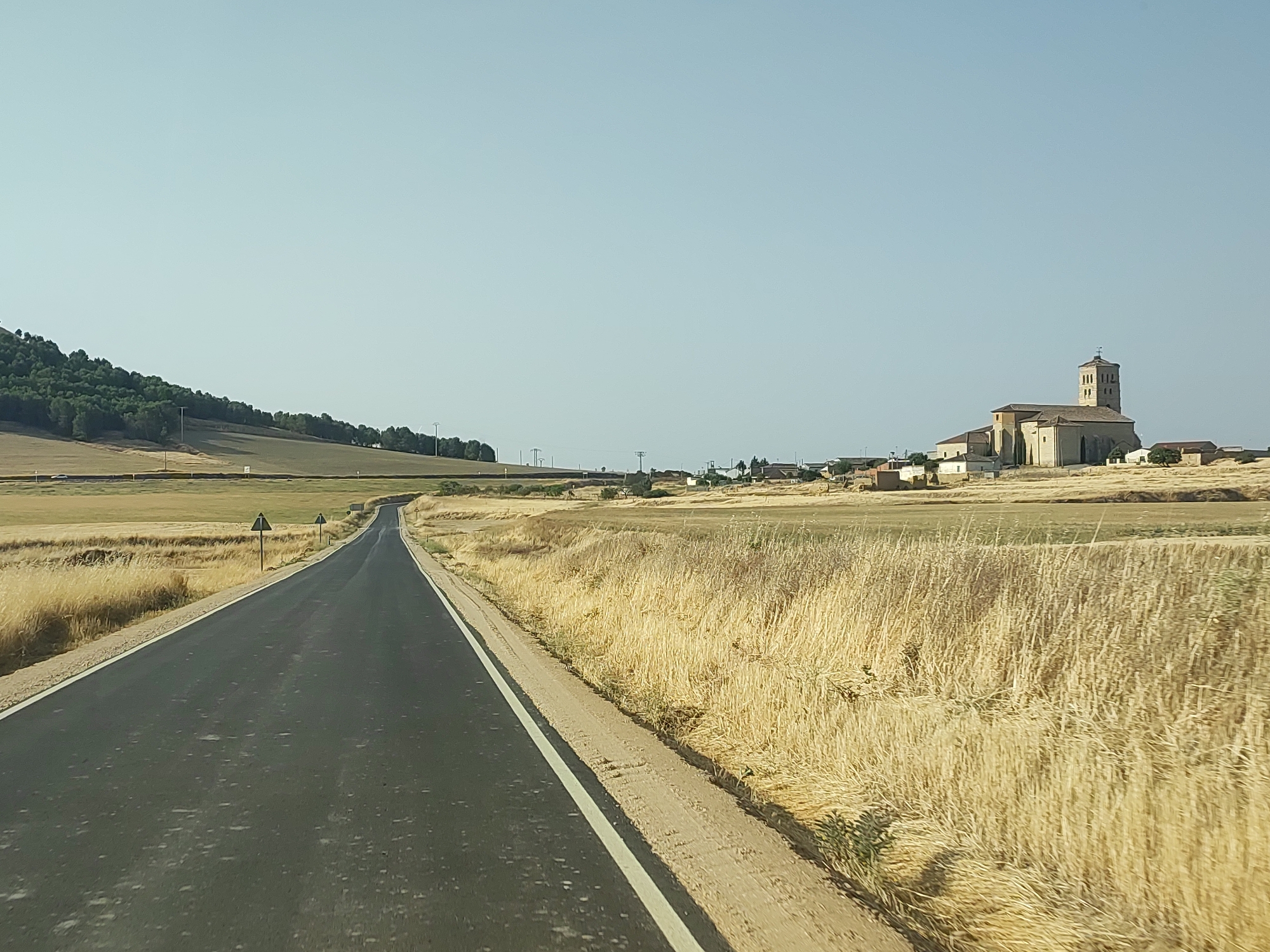 An asphalt road curves into the distance through a dry, rural landscape under a clear, pale blue sky. On the right side of the road, tall, golden-brown grass grows densely. In the mid-ground, a collection of buildings, including a prominent stone church with a bell tower, is visible in the distance. The church has a red-tiled roof and a cross at its apex. Further to the left, a treed hill rises. Two triangular warning signs are positioned along the side of the road. The scene appears to be a roadside view within or near the town of Torremormojón, Spain, characterized by its agricultural surroundings and a prominent church.