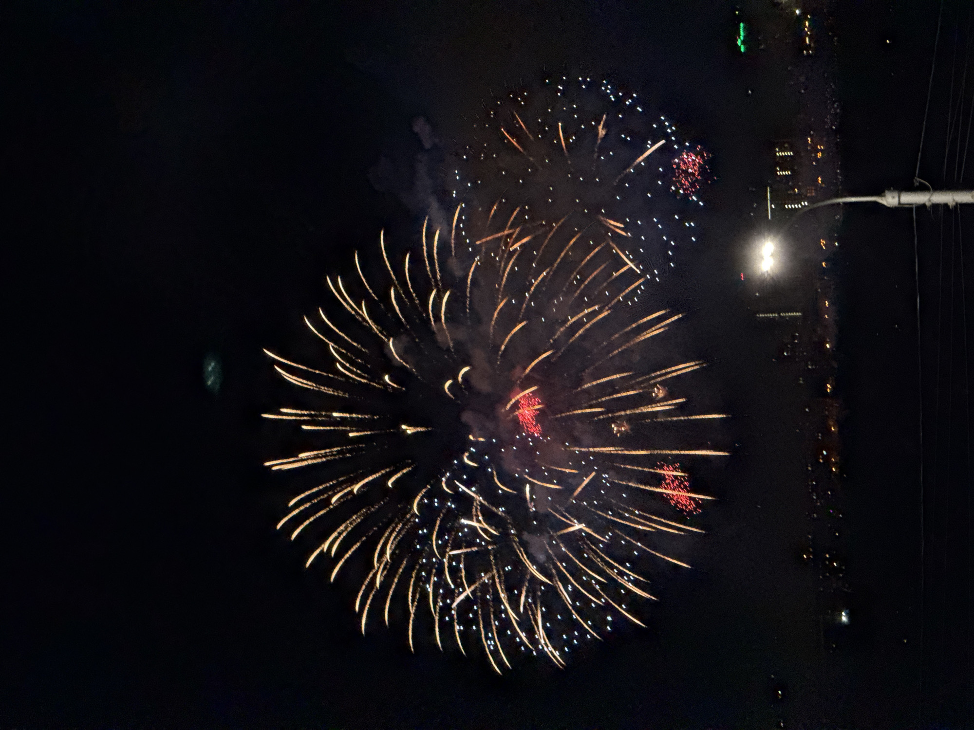 The image shows a nighttime view of fireworks exploding in the sky over a city. Two large bursts of fireworks dominate the scene, with a larger one in the center and a smaller one above and to the right. The fireworks display a variety of colors, including yellow, white, and red. In the background, city lights are visible along a waterfront. A lamppost with a bright light is visible in the right side of the frame. The location appears to be Krasnoyarsk, Russia, given the cityscape along the Yenisey River. The Yenisey River is visible in the background, with lights reflecting off the water. A pole is seen on the right of the photo.