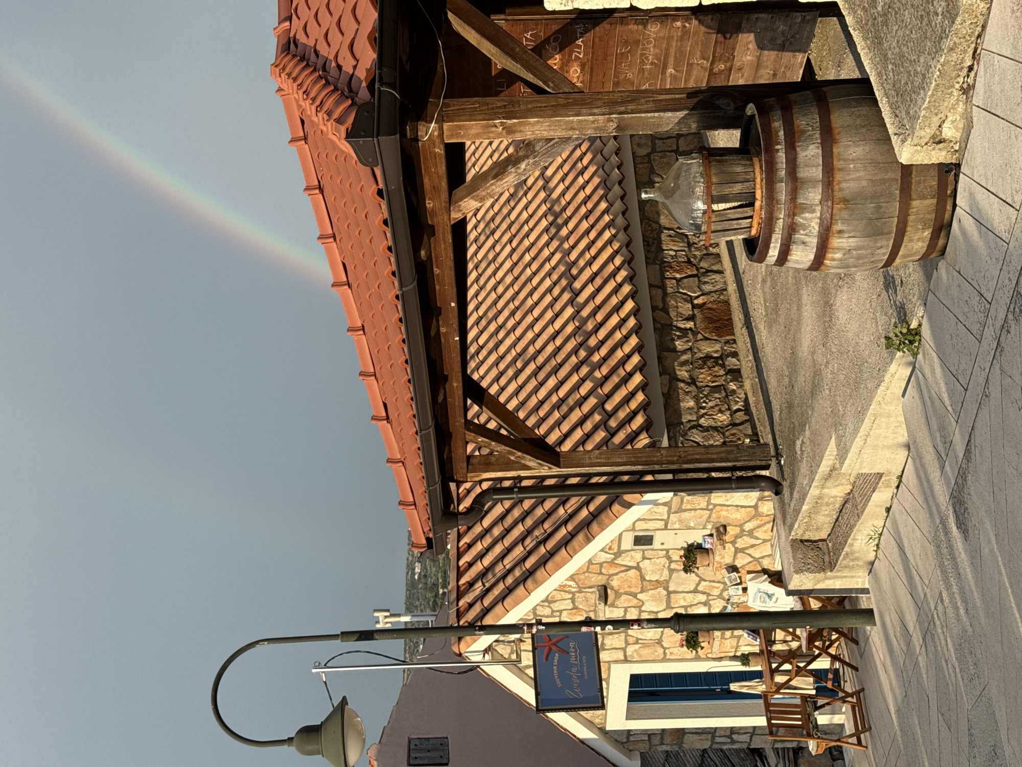 A low-angle, eye-level shot shows a quaint scene in Primošten, Croatia, with a vibrant rainbow arcing across a pale blue sky in the upper right. The foreground features a cobblestone pathway leading towards a stone building with a terracotta tiled roof. To the left, a small souvenir shop with a sign displaying a red starfish and the text "Zvizda mora" is visible, with a blue door and a small table and chairs set outside. A vintage street lamp stands to the left of the shop. A wooden barrel with a glass bottle on top sits to the right, in front of a wooden-covered area with a tiled roof. The building's exterior is made of rough-hewn stone, and a downspout runs along the right side of the structure. In the distance, a hint of the town and hills is visible. The lighting suggests late afternoon sun, casting long shadows on the ground.