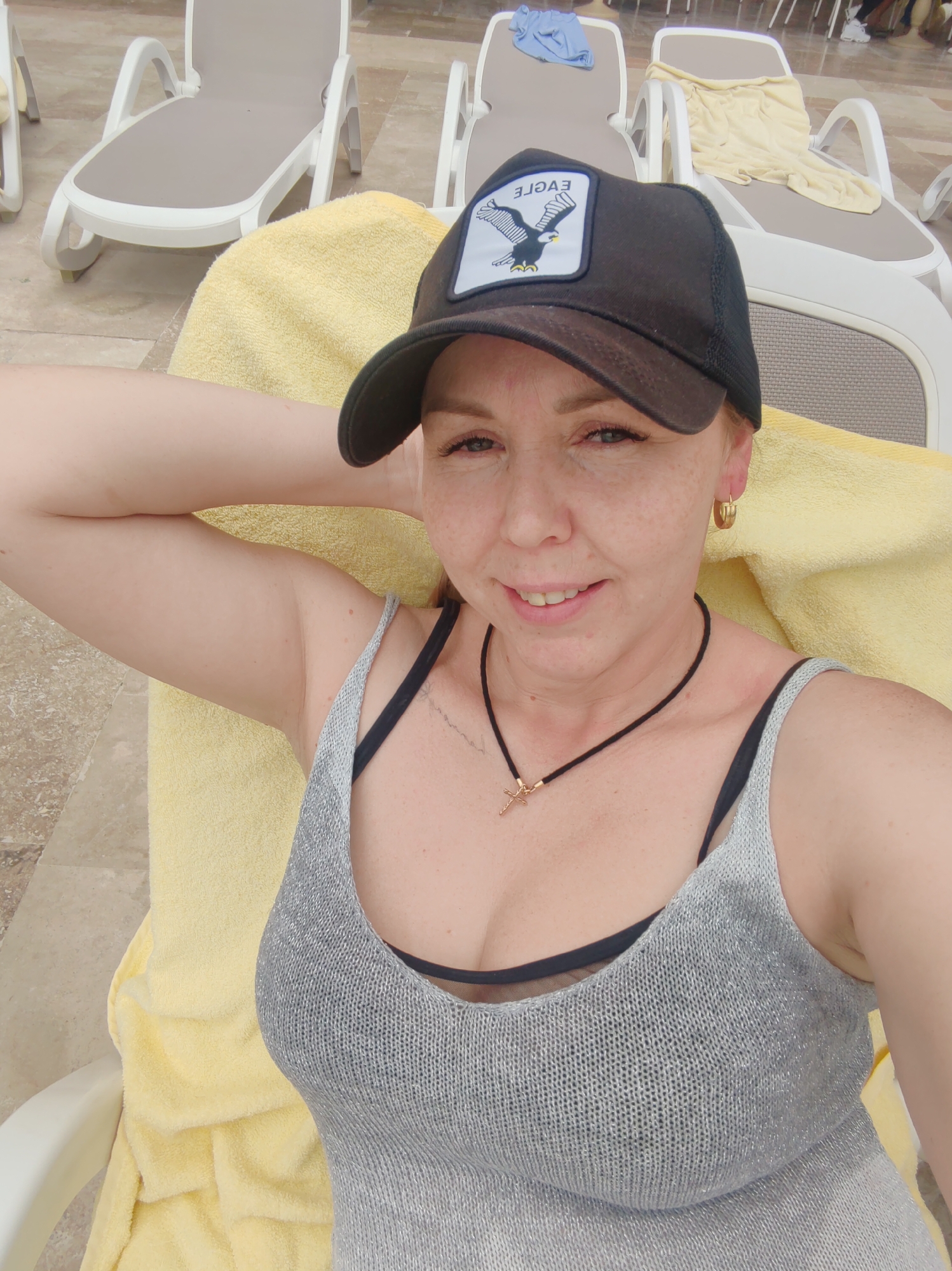 A woman is lying on a white chaise lounge. She is wearing a black "EAGLE" baseball cap, a grey tank top over a black bra, and a black necklace with a gold cross pendant. A yellow towel is beneath her. A pair of gold hoop earrings adorn her left ear. Several other chaise lounges can be seen in the background. The location is the city of Alanya, Turkey.