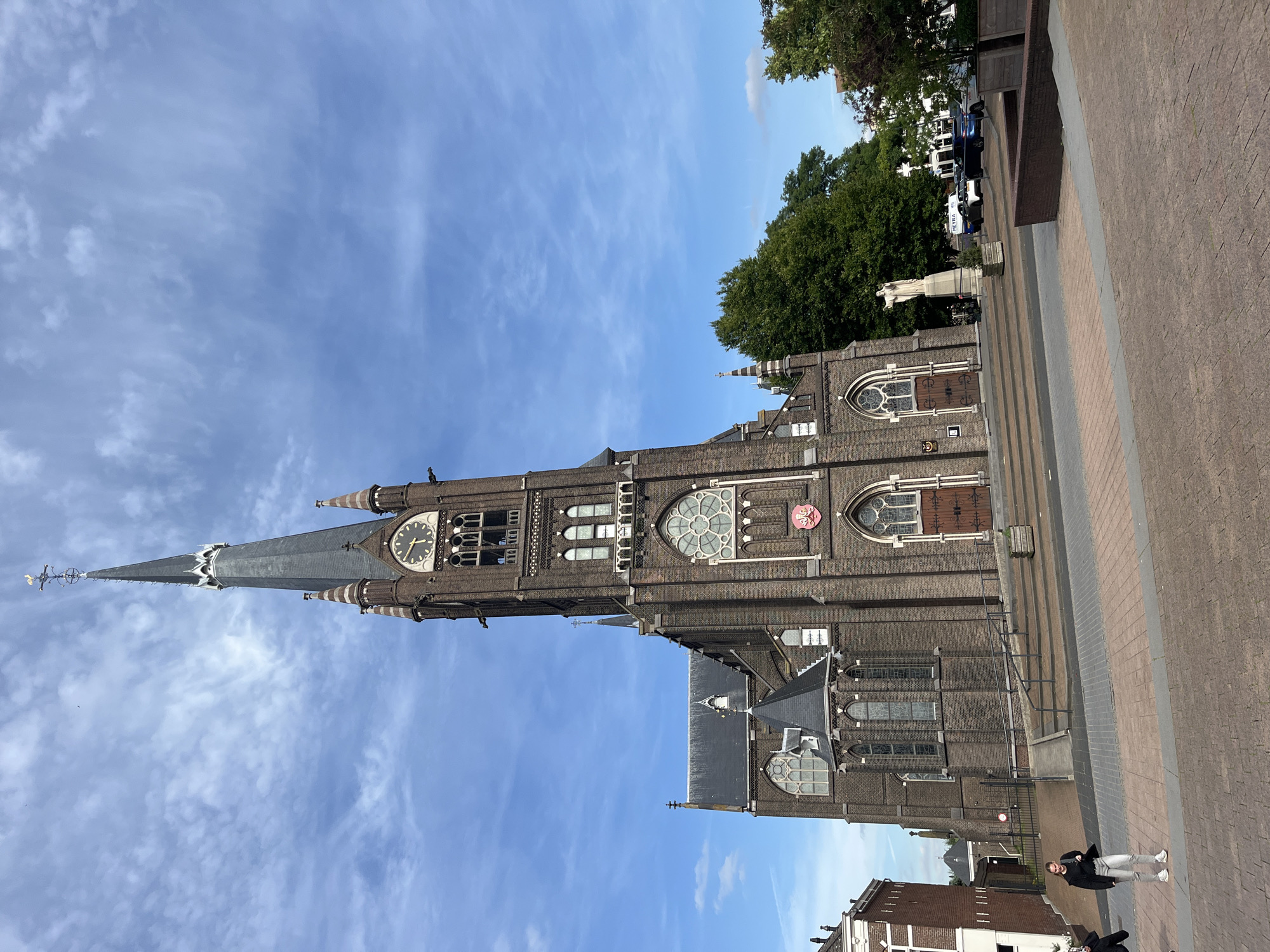 A full, eye-level, medium shot shows a large brick church with a tall, pointed spire against a partly cloudy blue sky. The spire is topped with a weather vane shaped like a rooster. The church has a clock on its tower, and ornate Gothic-style windows. The church is located in a city square with brick paving and a few trees. A white statue of a person stands in front of the church, and several cars are parked in the background. Two people are walking in the foreground.