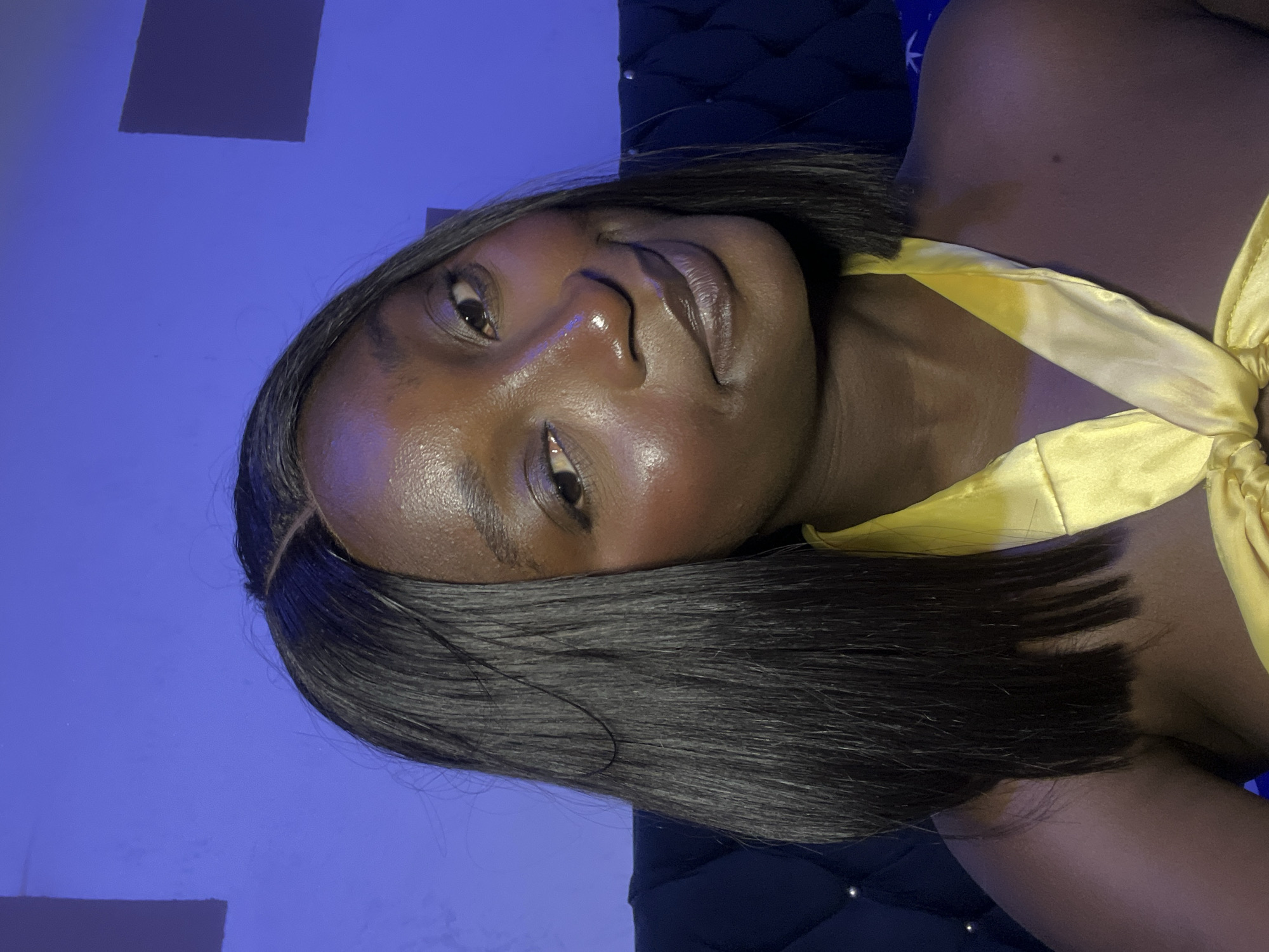 A close-up, slightly tilted, portrait of a dark-skinned woman with short, straight black hair. She has smooth skin, brown eyes, and is wearing a yellow satin top. The background is a combination of a light blue wall and a dark blue, textured surface, possibly a headboard. The location is presumably Jos, Nigeria.