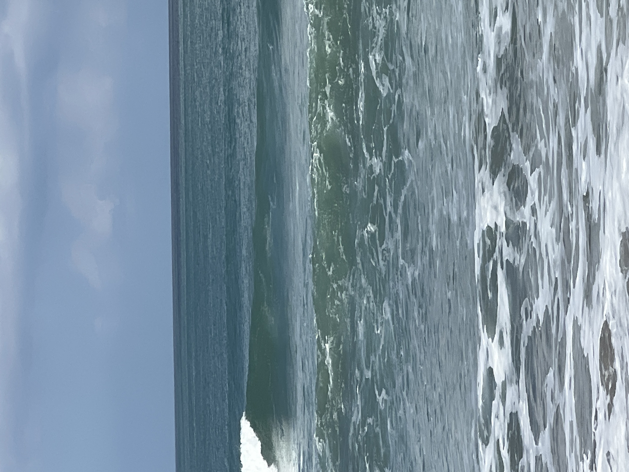 A medium shot captures the turbulent ocean waters off the coast of Kuta, Indonesia. The scene is dominated by rolling waves, with the closest wave breaking into white foam. The water is a deep teal color, reflecting the overcast sky above. The horizon line is a clear, straight division between the water and the pale blue sky, which is streaked with soft, grey clouds. The overall impression is one of natural power and dynamic movement. No people or man-made objects are visible in the frame.