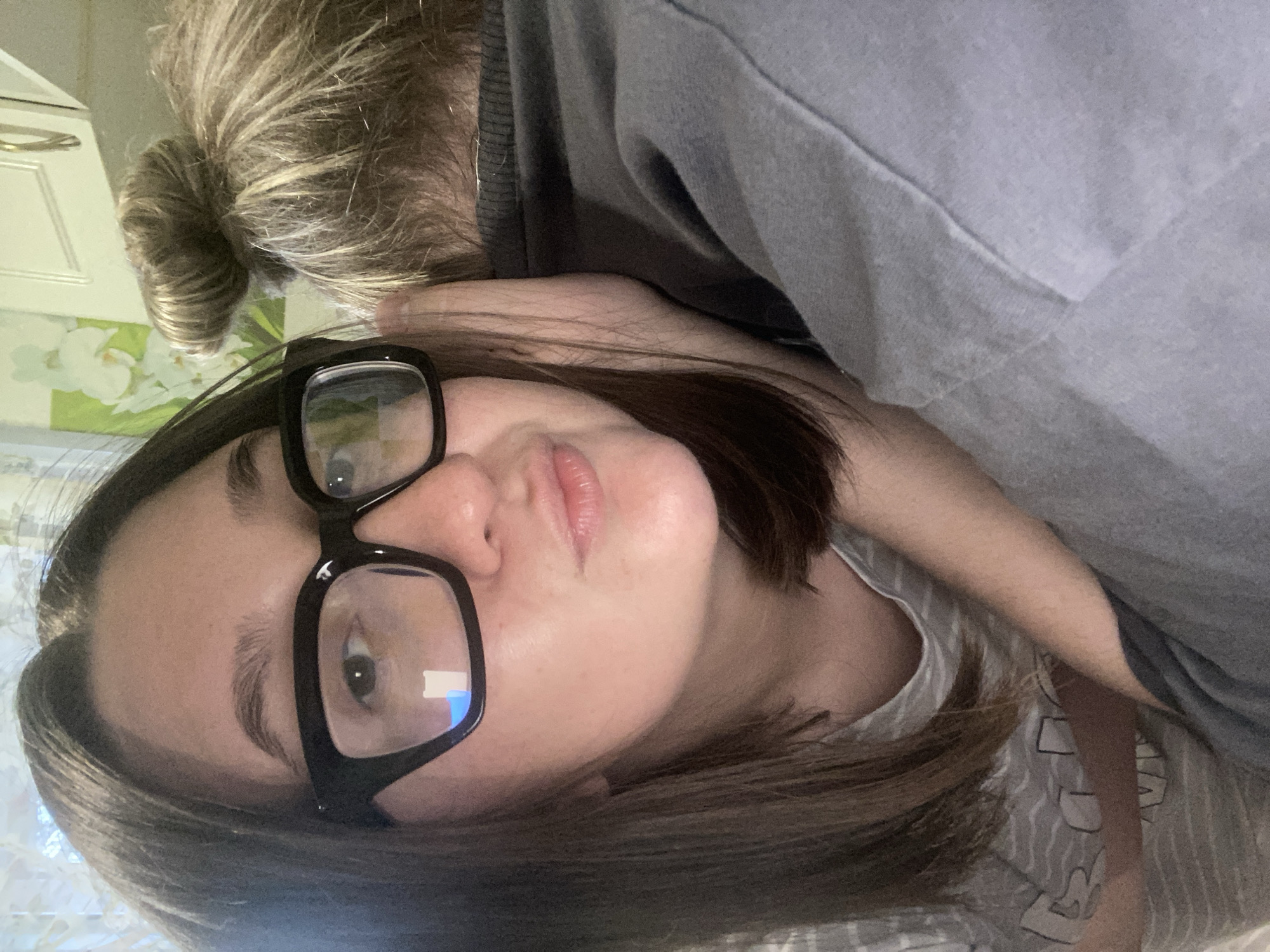A selfie of a woman indoors. She has shoulder-length brown hair, wears black glasses, and has fair skin. She is wearing a gray shirt and a gray sweatshirt is draped over her. Part of another person's bun is visible in the top left corner. There is a white cabinet and a floral pattern in the background. The location is potentially Chaikovsky, Russia.