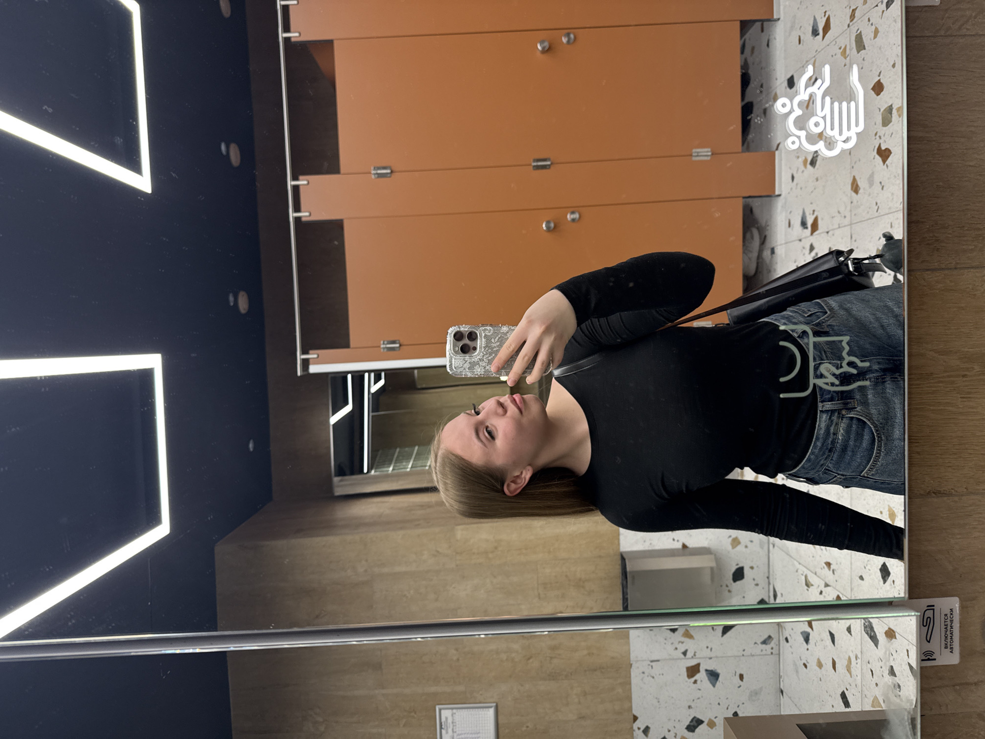 A young adult female is depicted taking a mirror selfie in a public restroom located in the city of Sochi, Russia. The female, with long straight light brown hair, is wearing a black long-sleeved top, blue denim jeans, and a light-colored belt with an ornate buckle. She holds a smartphone encased in a patterned, possibly clear, cover in her right hand, while looking to her left. The restroom interior, visible in the reflection, features multiple brown-orange colored stall doors with silver-toned hardware (hinges, top rails). The walls are finished in a light brown, possibly textured, material. The floor is a light-colored terrazzo with irregular dark brown, black, and bluish-gray aggregate chips. The ceiling is dark, likely black, with prominent rectangular linear LED light fixtures and smaller circular recessed lights. A wall-mounted fixture, possibly a hand dryer, is partially visible on the left side of the reflection. On the lower left of the mirror, a white decal shows a hand under a faucet with the Russian text "ВКЛЮЧАЕТСЯ АВТОМАТИЧЕСКИ" (Turns on automatically) below it. On the lower right of the mirror, a white graphic symbol depicting handwashing with suds is present.