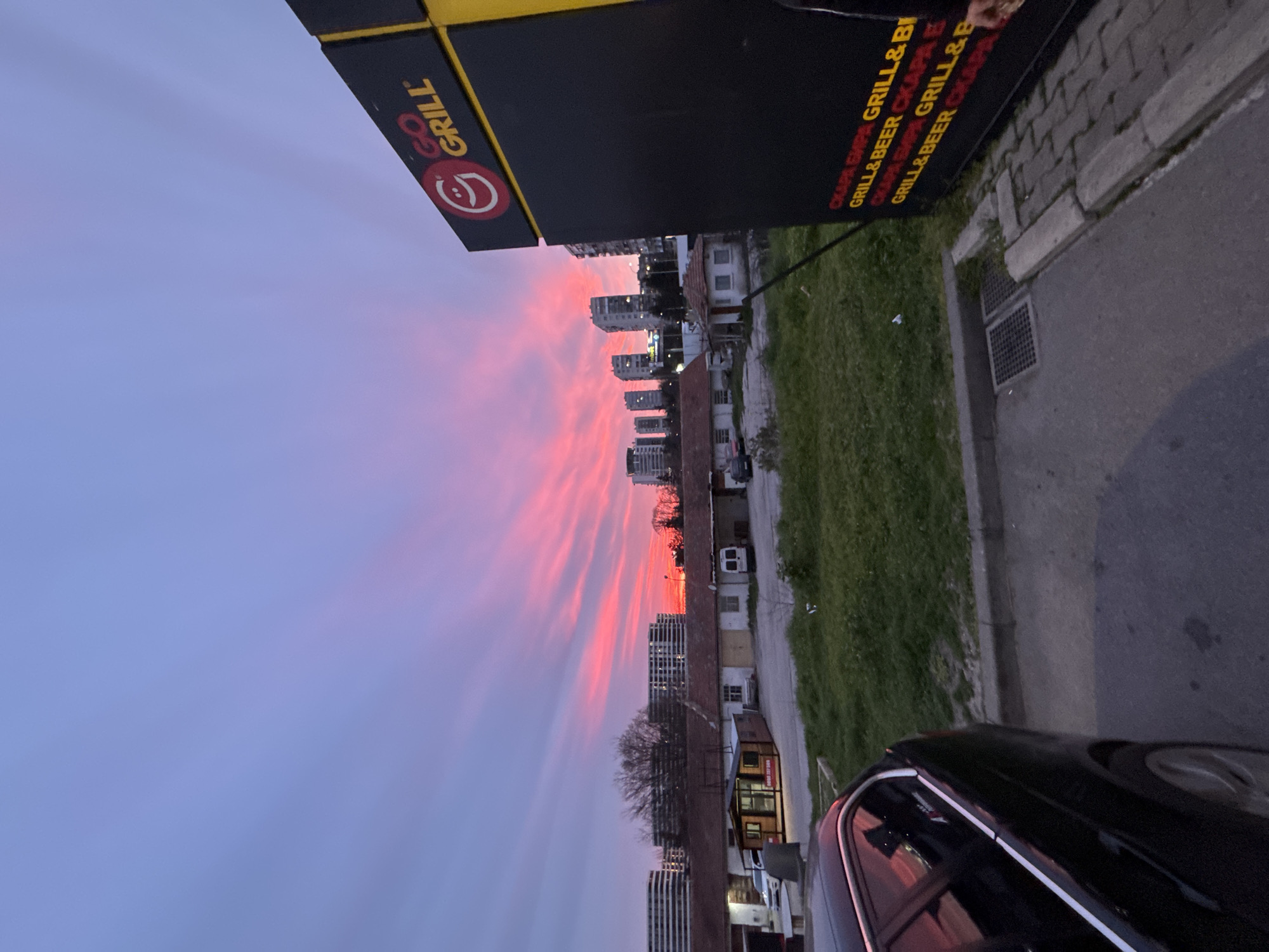 A dusk cityscape in Varna, Bulgaria, is captured at a skewed angle, displaying the sky painted in vivid pink and violet hues. A 'GO GRILL' restaurant's sign is prominent on the upper left, next to another sign repeating "СКАРА БИРА GRILL&B GRILL&BEER". Buildings, ranging from houses to modern skyscrapers, line the horizon, set against the dramatic sky. A patch of greenery runs parallel to a paved street, with a black car visible at the bottom right. A sewer grate lies on the pavement.