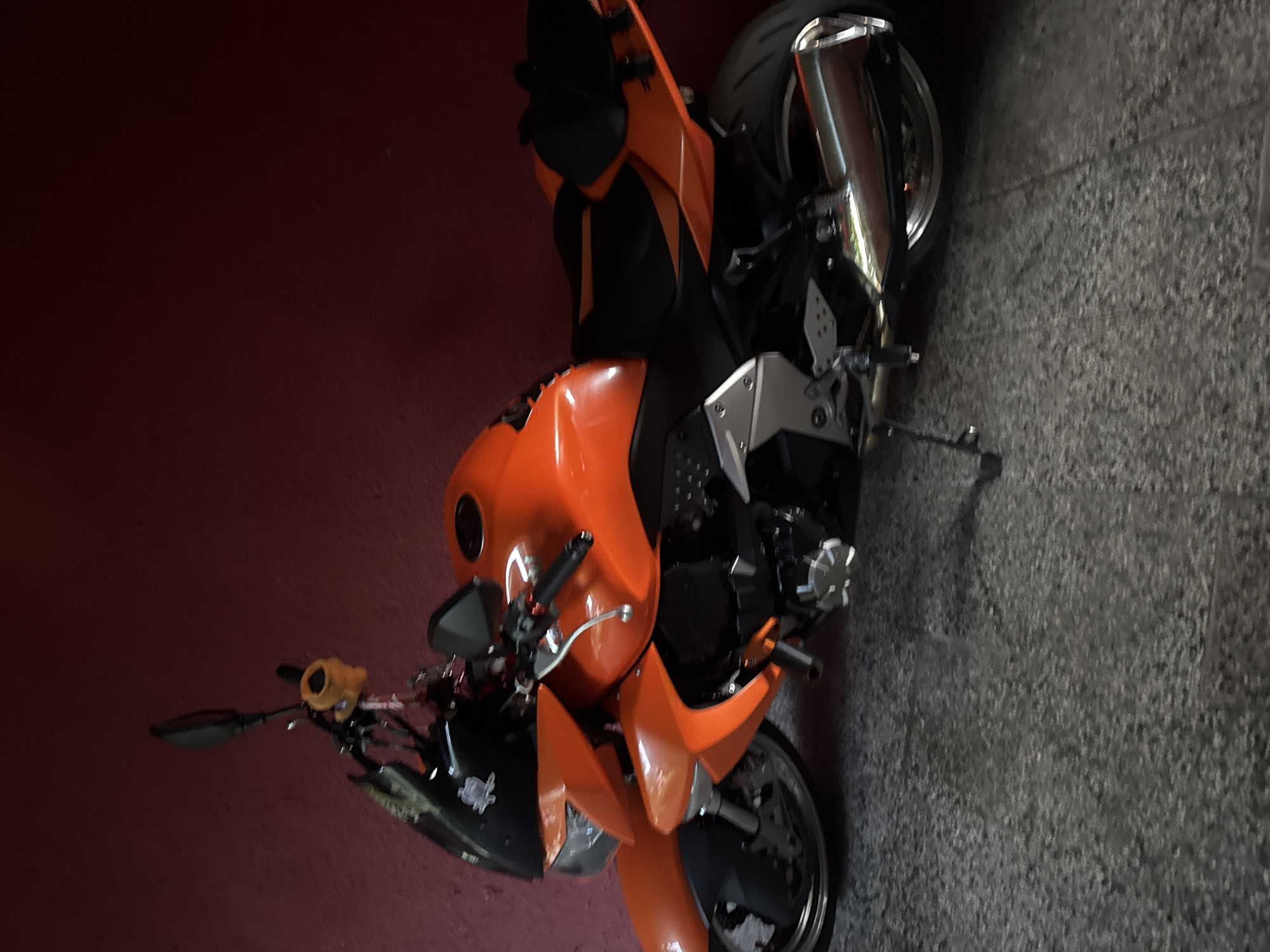 A bright orange and black motorcycle is depicted parked against a deep reddish-pink wall on a mottled gray concrete floor in an unspecified indoor or sheltered location within the city of Unknown, Unknown. The motorcycle is oriented to the left, resting on its deployed kickstand. Key visible components include a chrome exhaust pipe on the right side, black soft luggage attached to the rear seat, and a small yellowish-orange ornamental object suspended from the handlebars. No individuals are present in the frame. The scene is illuminated by subdued, ambient light.