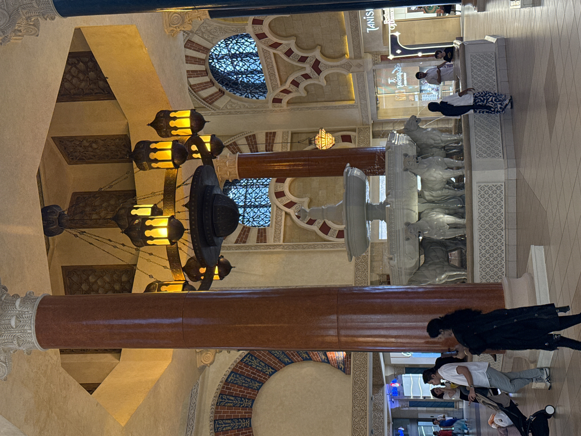 The image depicts an indoor atrium with distinct architectural elements reminiscent of Islamic design. Dominating the center is a large, ornate chandelier with multiple hanging lamps that emit a warm, yellow light. Flanking the chandelier are tall, reddish-brown pillars with detailed white capitals. The walls feature arched doorways and windows with intricate geometric latticework, along with decorative brickwork and patterned friezes.
In the foreground, a white marble fountain is visible, adorned with sculptures of what appear to be lions or similar animals. Several people are present in the scene. On the left, a man in a white shirt and grey pants pushes a stroller, accompanied by a woman in a black outfit. Further back, a woman wearing a black headscarf and a patterned skirt stands near the fountain. To the right of the fountain, a man in a light-colored shirt and a child are standing, seemingly interacting with the fountain area. In the background, storefronts are visible, one displaying the signage "TANISHQ" and "Festival of Diamonds". The overall environment suggests a spacious, well-lit indoor public space, likely a shopping mall or a similar establishment. Based on the architectural style and the presence of known retail brands, the location is likely a high-end commercial area in Dubai, United Arab Emirates.