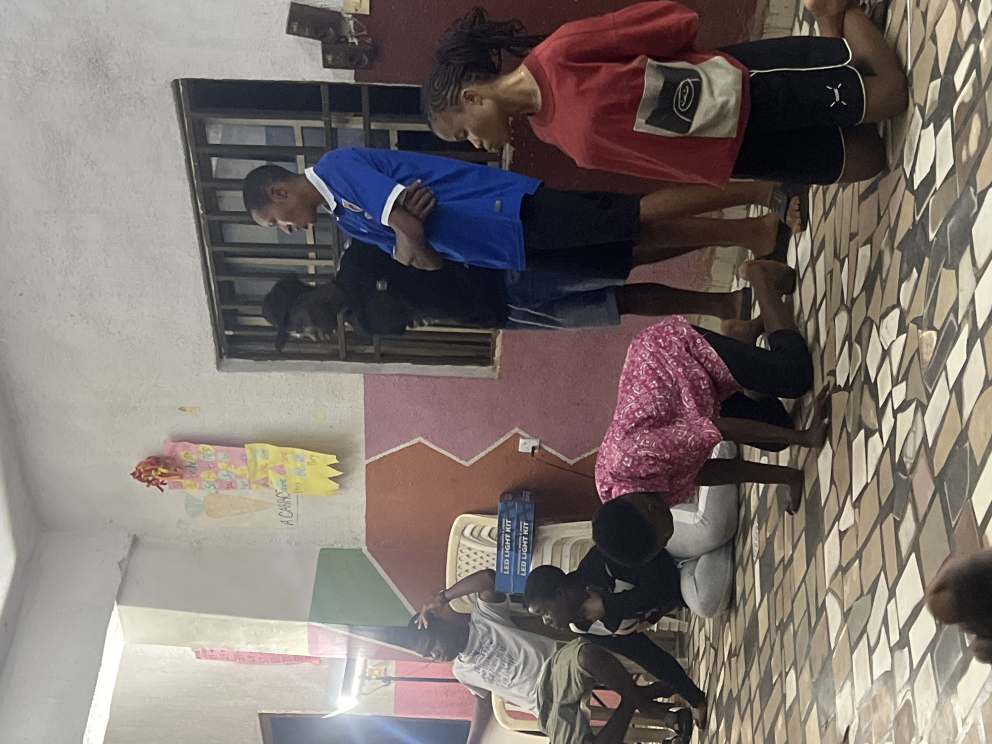 In Nkpor, Nigeria, a group of young people are gathered in a room with patterned tiled flooring. A young woman in a red long-sleeved shirt and black shorts is kneeling, facing away from the camera. Another young person, wearing a pink patterned shirt and dark pants, is on their hands and knees on the floor, also facing away from the camera. To their left, a person in a black shirt and grey pants sits on a chair, looking upwards. To the right of the person on the chair, two young men stand. One is wearing a black t-shirt and blue shorts, and the other is wearing a blue t-shirt with white trim and black shorts. They are looking towards something out of frame. The walls of the room are painted in bold geometric patterns of pink, orange, and green. Two boxes labeled "LED LIGHT KIT" are stacked on chairs in the background. A window with metal bars is visible on the far wall, and posters are displayed on the wall to the left. The lighting suggests it is daytime, with a bright light source to the far left.