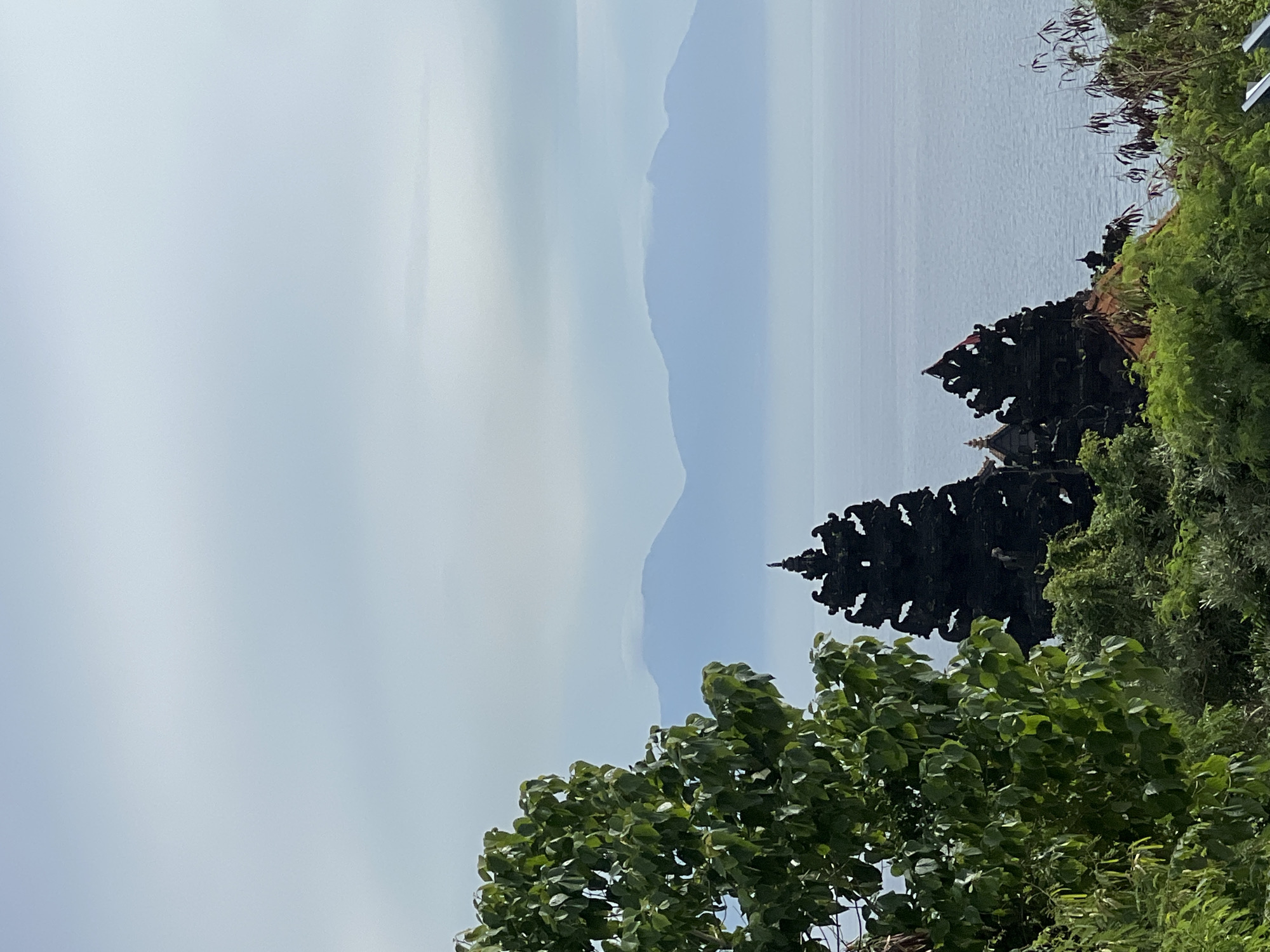 The image captures a coastal view in Kuta Selatan, Indonesia, likely the Uluwatu Temple area. A black silhouette of a tiered Balinese temple stands on a cliff edge amidst lush green vegetation. The ocean stretches out to the horizon with distant, indistinct mountain outlines under a hazy sky. The sky occupies the upper left of the image. The foreground is dominated by trees and foliage.