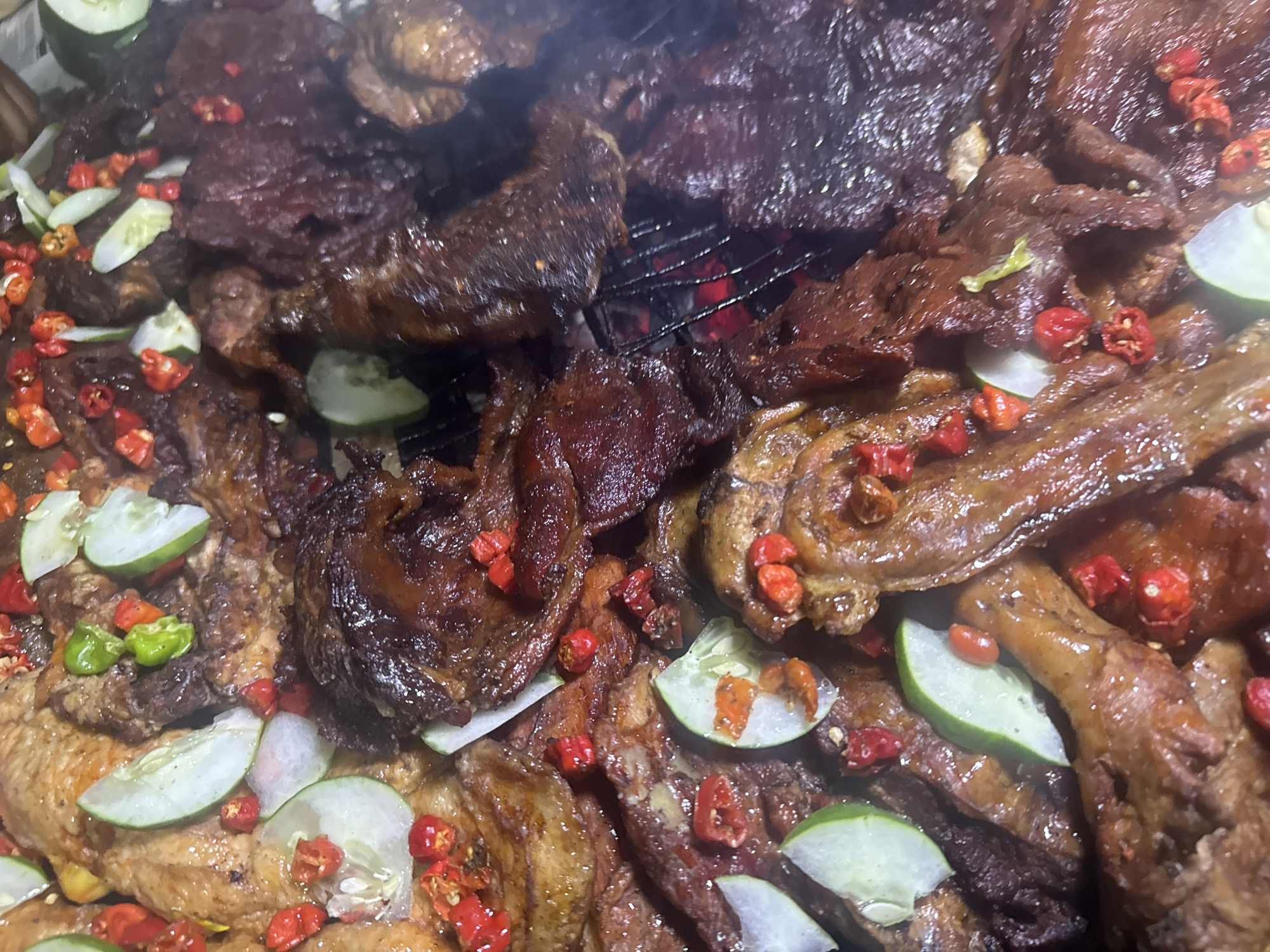 A close-up, overhead shot captures a platter of grilled meat, adorned with slices of cucumber and small red chili peppers. The meat, appearing dark brown and glistening, is arranged in a dense, overlapping fashion. Some pieces show char marks, suggesting they've been cooked over an open flame, with hints of red embers visible beneath the meat in the lower right quadrant. The cucumber slices are thinly cut and round, interspersed with the vibrant red of the chilies, creating a contrast in color and texture. A few small green chili pieces are also scattered among the ingredients. The overall impression is one of a flavorful, spicy dish, likely prepared as street food. Given the description of grilled meats with these specific garnishes, this dish is highly likely to be found at a roadside food stall or a local eatery in the city of Abuja, Nigeria.