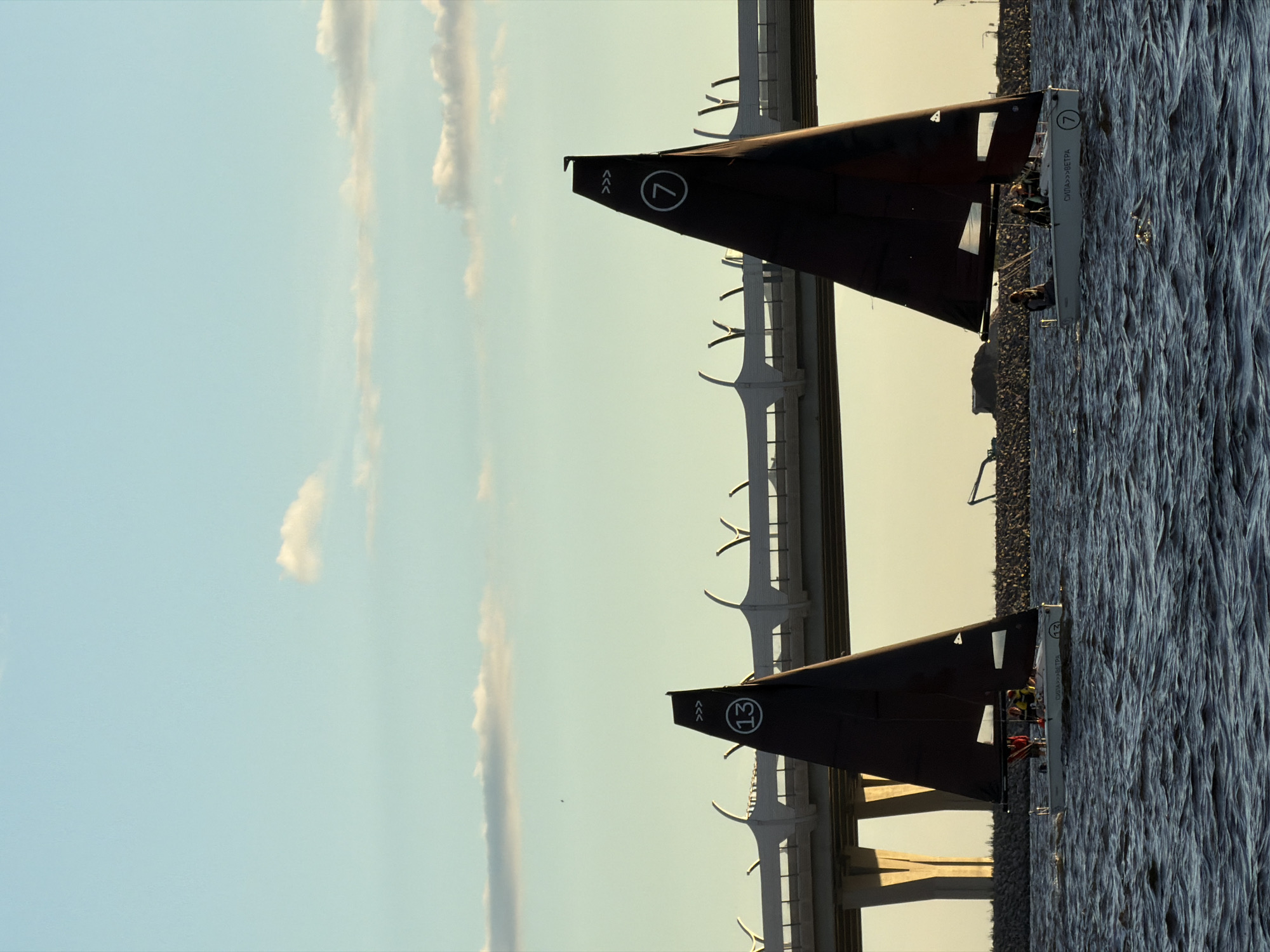 The image depicts a body of water, likely a river or gulf, with two sailboats under a pale blue sky with scattered clouds. In the background, a modern bridge with a distinctive white, curved railing stretches across the scene. The water is choppy, indicating a moderate breeze.
The sailboat on the left has a dark triangular sail with a white circle containing the number "13" and some directional arrows. The sailboat on the right also has a dark triangular sail, with a white circle containing the number "7" and similar directional arrows. Both sailboats have a white hull. The sailboat on the right has several individuals on board, some wearing life vests. Text is visible on the hull of the sailboat on the right, reading "СИЛА >>> ВЕТРА" (Sila >>> Vetra), which translates to "Force >>> Wind".
The overall setting suggests a recreational sailing activity, potentially near an urban area given the prominent bridge structure. This bridge is identifiable as the Western High-Speed Diameter in Saint Petersburg, Russia, a major transportation artery connecting different parts of the city and its suburbs. The presence of the bridge and the open water context strongly point to this location. The lighting suggests it is either early morning or late afternoon.