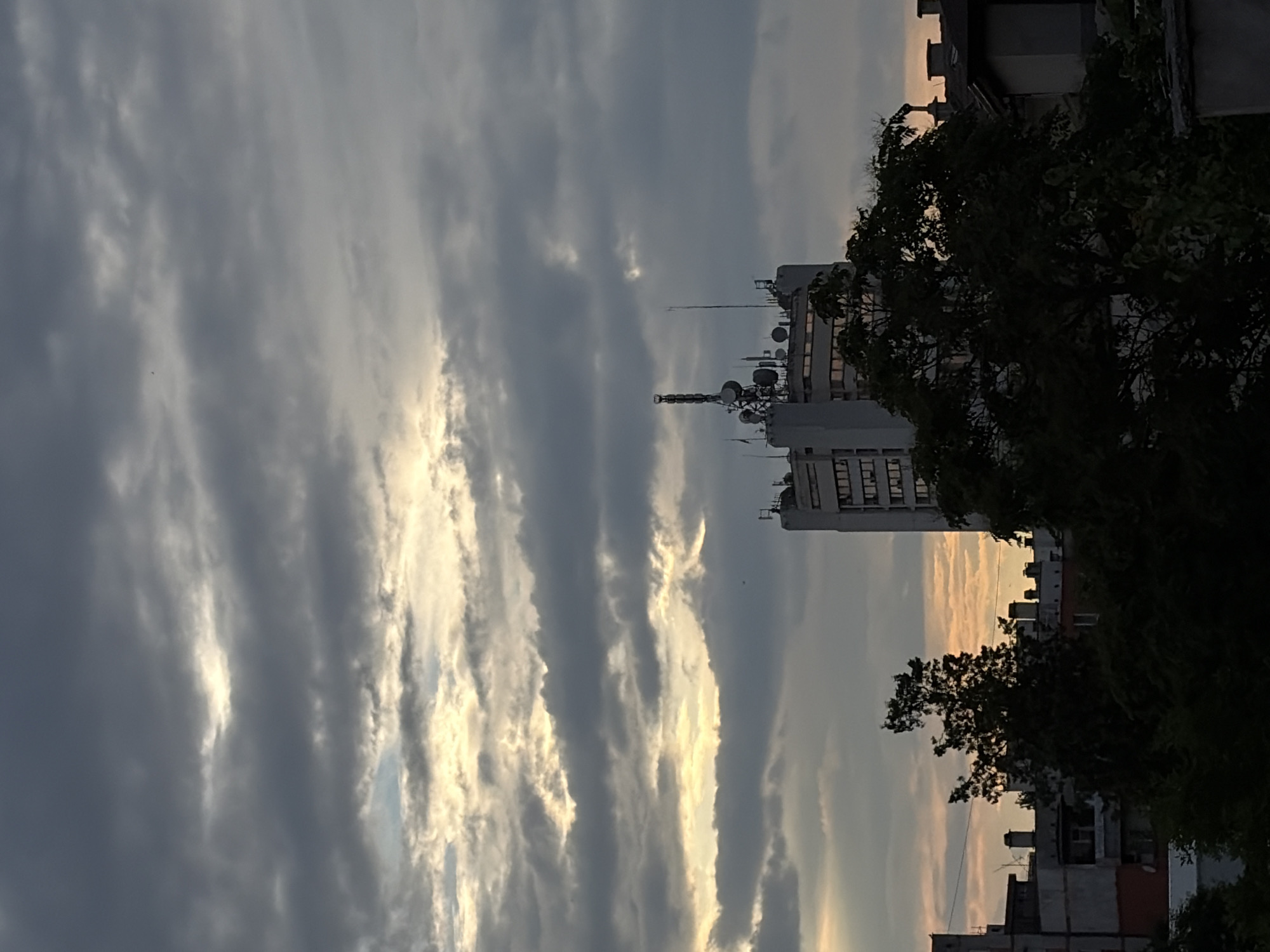This is a landscape photograph with a vertical orientation, featuring a cloudy sky and urban buildings. The scene is dominated by a dramatic sky with layered clouds, some appearing dark and stormy, while others are illuminated by a soft, golden light, suggesting either sunrise or sunset. In the mid-ground, a tall, rectangular building with multiple antennas and satellite dishes on its rooftop stands prominently. This building appears to be an office or communication tower. To the right, a large, leafy tree partially obscures the view of other buildings, which are more residential in appearance, with visible windows and balconies. The overall atmosphere is somewhat somber due to the heavy cloud cover, but the warm light breaking through adds a touch of beauty. The image seems to capture a moment in an urban environment, possibly a city, during twilight. There is no visible text in the image.