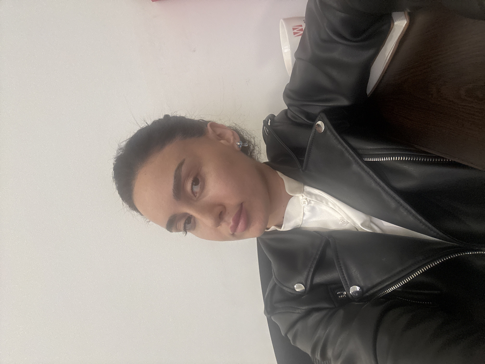 A woman with dark hair pulled back is wearing a black leather jacket over a white collared shirt. She has fair skin and is wearing small earrings. The background is a plain white wall with a dark wooden surface visible in the upper right corner. The location is not discernible from the image.