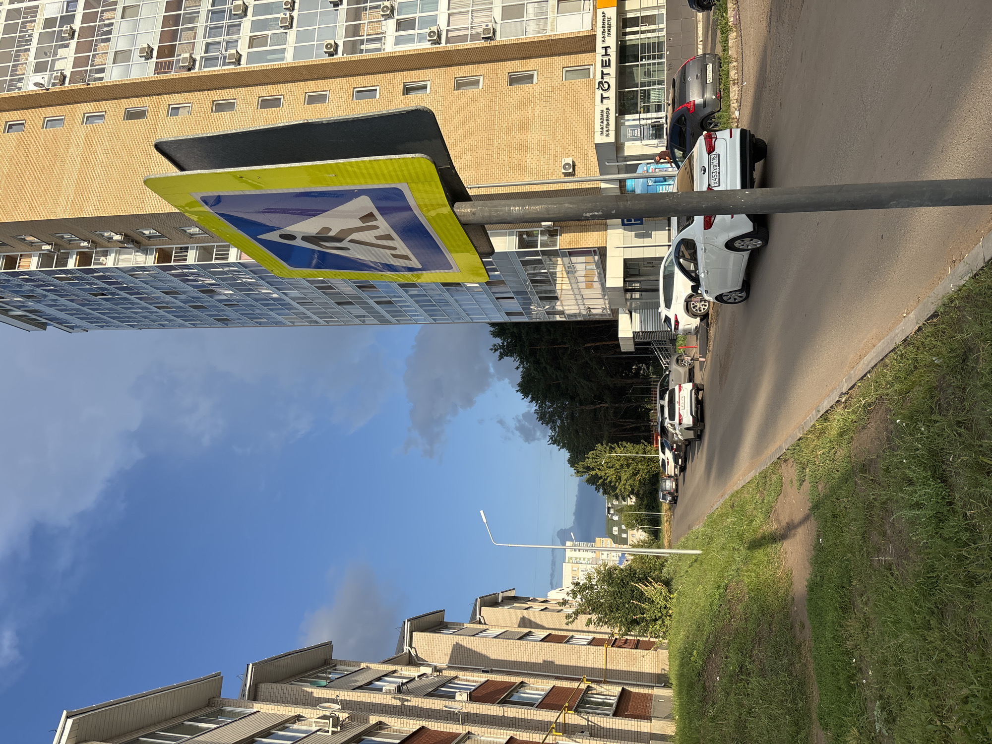 The image shows a street scene in the city of Naberezhnye Chelny, Russia, on a sunny day with scattered clouds. In the foreground, a pedestrian crossing sign stands on a pole. To the left, a light-colored multi-story residential building runs along the street. To the right, a tall, modern brick building with many windows and air conditioning units is visible. Several cars are parked or driving on the asphalt road. Green grass and trees line the side of the road and the median. The overall impression is of a typical urban residential area.