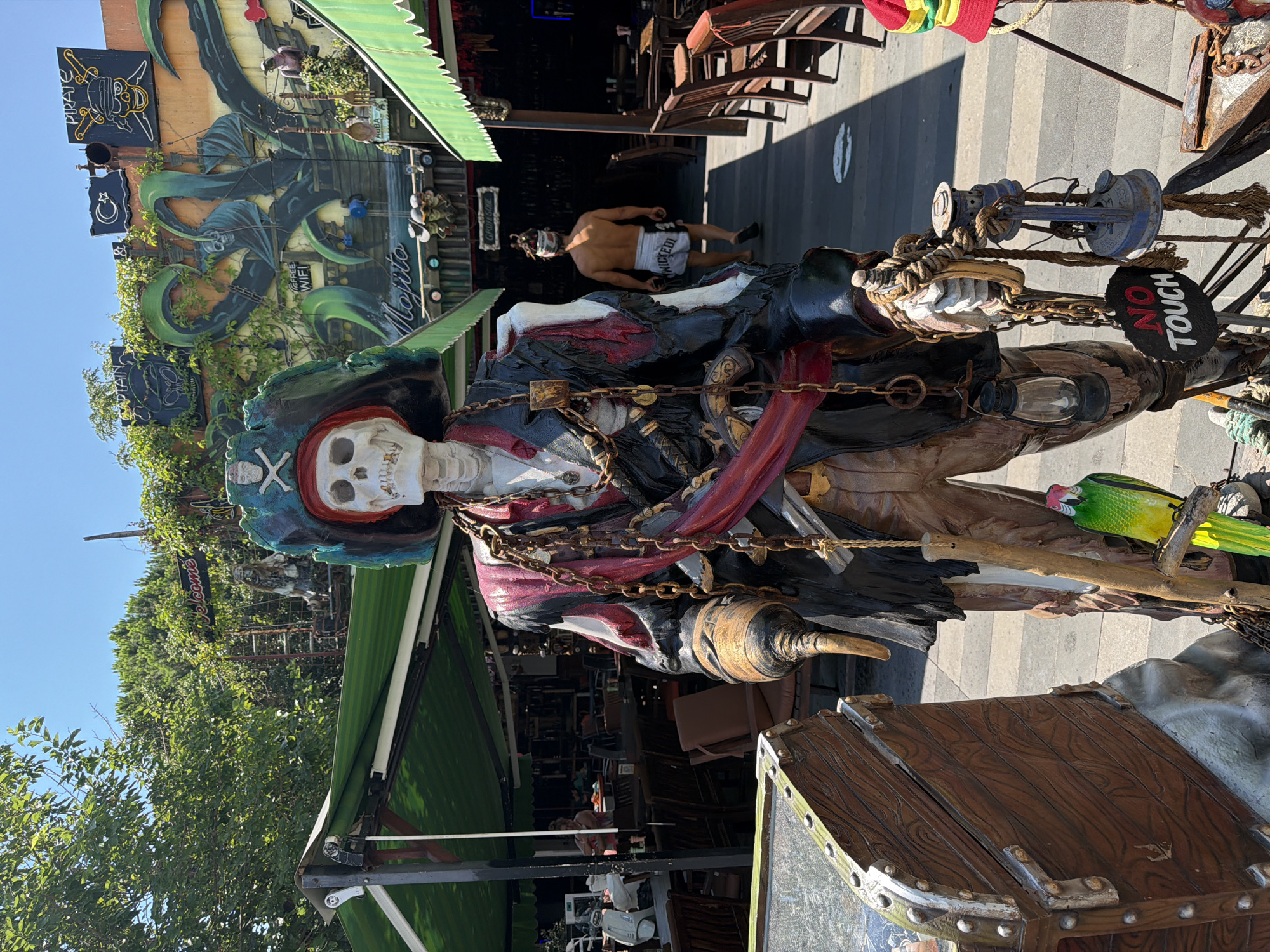 The image shows a life-sized pirate skeleton statue with a parrot on its shoulder, standing in front of a building decorated with pirate-themed murals and signs. The building is likely a bar or restaurant in Kemer, Turkey, given the "Mojito" sign and the overall vibrant atmosphere. The statue is adorned with pirate regalia, including a tricorn hat, chains, and a sash. A wooden treasure chest is placed in the foreground to the left of the statue. A man with his back to the camera, wearing shorts and a headscarf, is walking away from the statue into the building. The scene appears to be an outdoor seating area, with tables and chairs visible in the background. The sky is clear and blue, suggesting a sunny day. The overall impression is a tourist-oriented establishment aiming for a fun, pirate-themed experience.