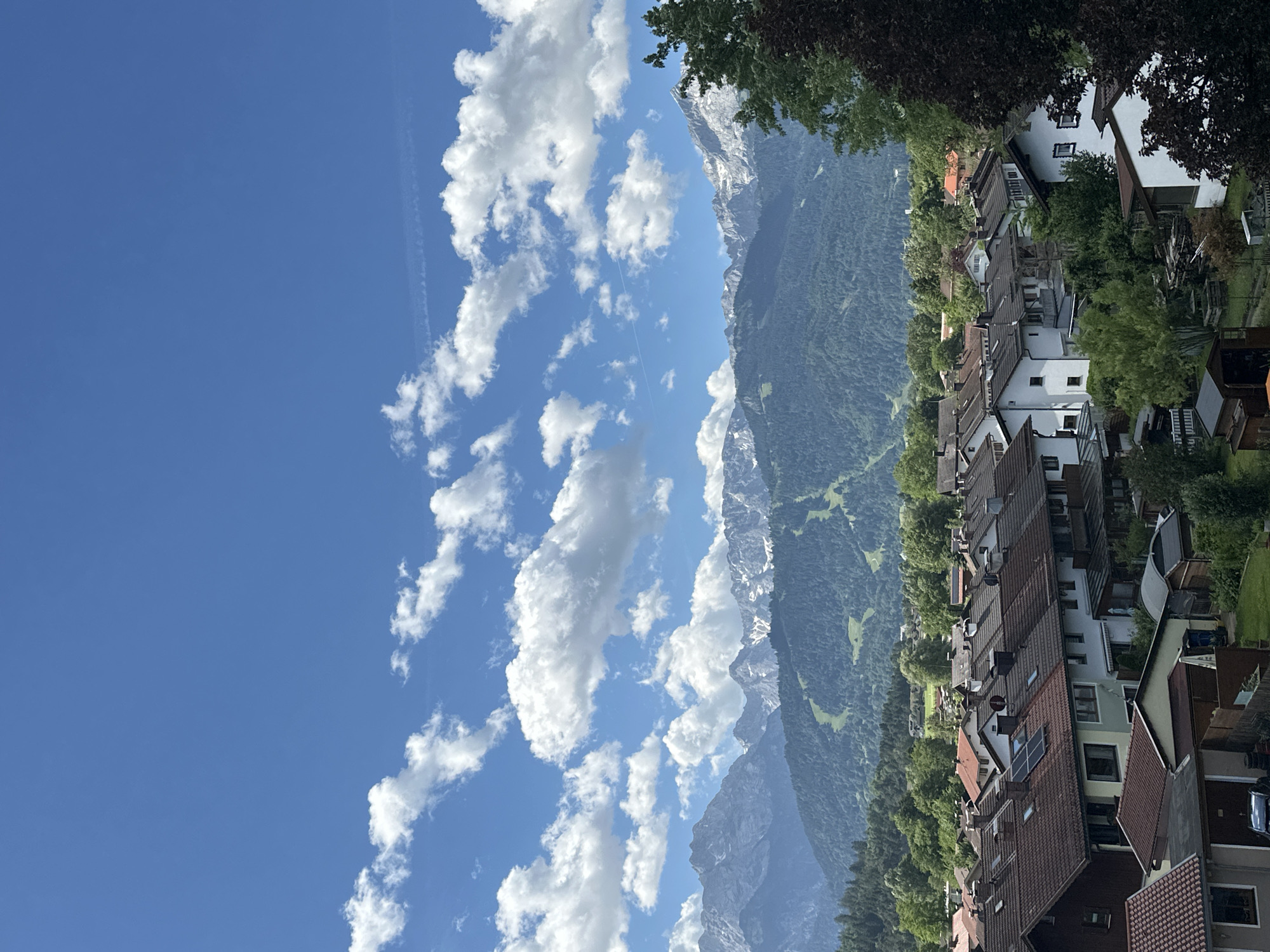 The image depicts a picturesque view of Garmisch-Partenkirchen, Germany, on a bright, sunny day. In the foreground, a dense cluster of Bavarian-style houses with characteristic dark tiled roofs fills the lower half of the frame. Many of these houses have white or light-colored walls and multiple stories. Some roofs feature solar panels, indicating a modern integration of renewable energy. Small yards and gardens are visible between the houses, with trees and some sheds.
The middle ground is dominated by rolling green hills covered in dense forest, with visible ski slopes etched into the mountainside, suggesting the area's dual role as a summer resort and winter sports destination.
In the background, a majestic mountain range, likely the Alps, forms a dramatic backdrop. The peaks are rugged and some are capped with snow, even in what appears to be a warmer season. The sky above is a vibrant blue, dotted with fluffy white cumulus clouds, some of which are casting soft shadows on the landscape below. A faint contrail from an airplane is visible in the upper right quadrant of the sky. The overall atmosphere is one of natural beauty and serene, well-maintained urban development nestled within a stunning alpine environment. There are no people or animals visible in the frame.
