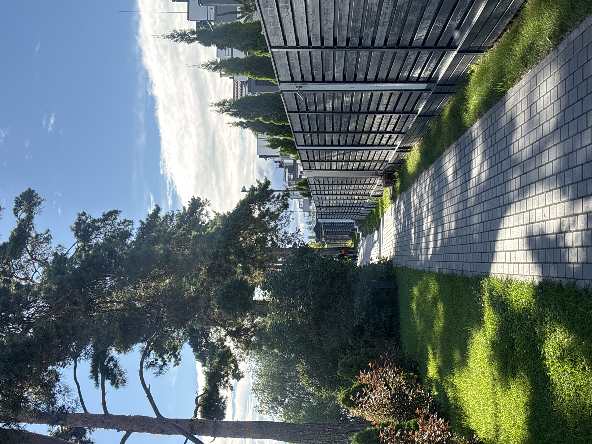 A paved walkway runs alongside a dark wooden fence, leading away from the viewer. Lush green grass lines the left side of the walkway, with ornamental bushes and large trees creating shade. The walkway is illuminated by dappled sunlight, casting geometric shadows. Two individuals are visible walking on the pathway in the distance, appearing as small figures. The fence, constructed of horizontal dark planks, encloses a property, and beyond it, modern residential buildings with green coniferous trees are visible. The sky above is a bright blue with scattered white clouds. The scene depicts a well-maintained residential area with a clear demarcation between public and private spaces.