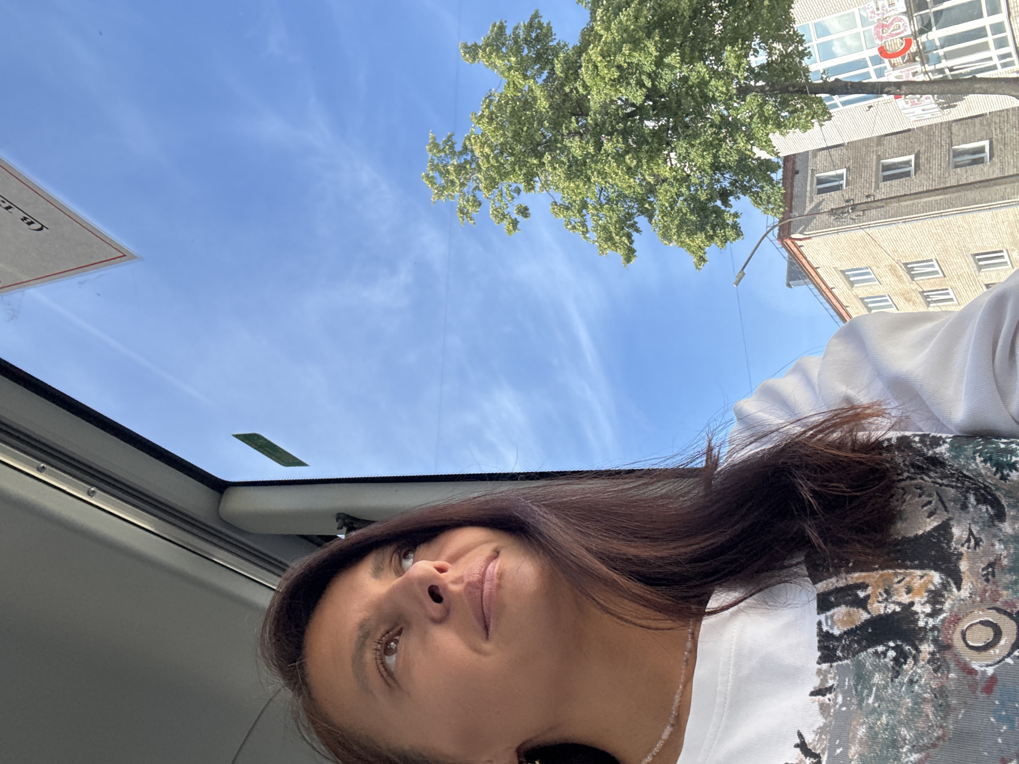A woman is seen from a low angle, looking up towards a clear blue sky with some wispy clouds. The edge of a car's sunroof frames the lower portion of the image, suggesting she is inside a vehicle. Above her, a tree with dense green foliage is visible against the sky. Beyond the tree, a building with multiple windows can be seen. The woman has dark hair and is wearing a white top. Based on the building design, this photo may have been taken in Chaikovsky, Russia.