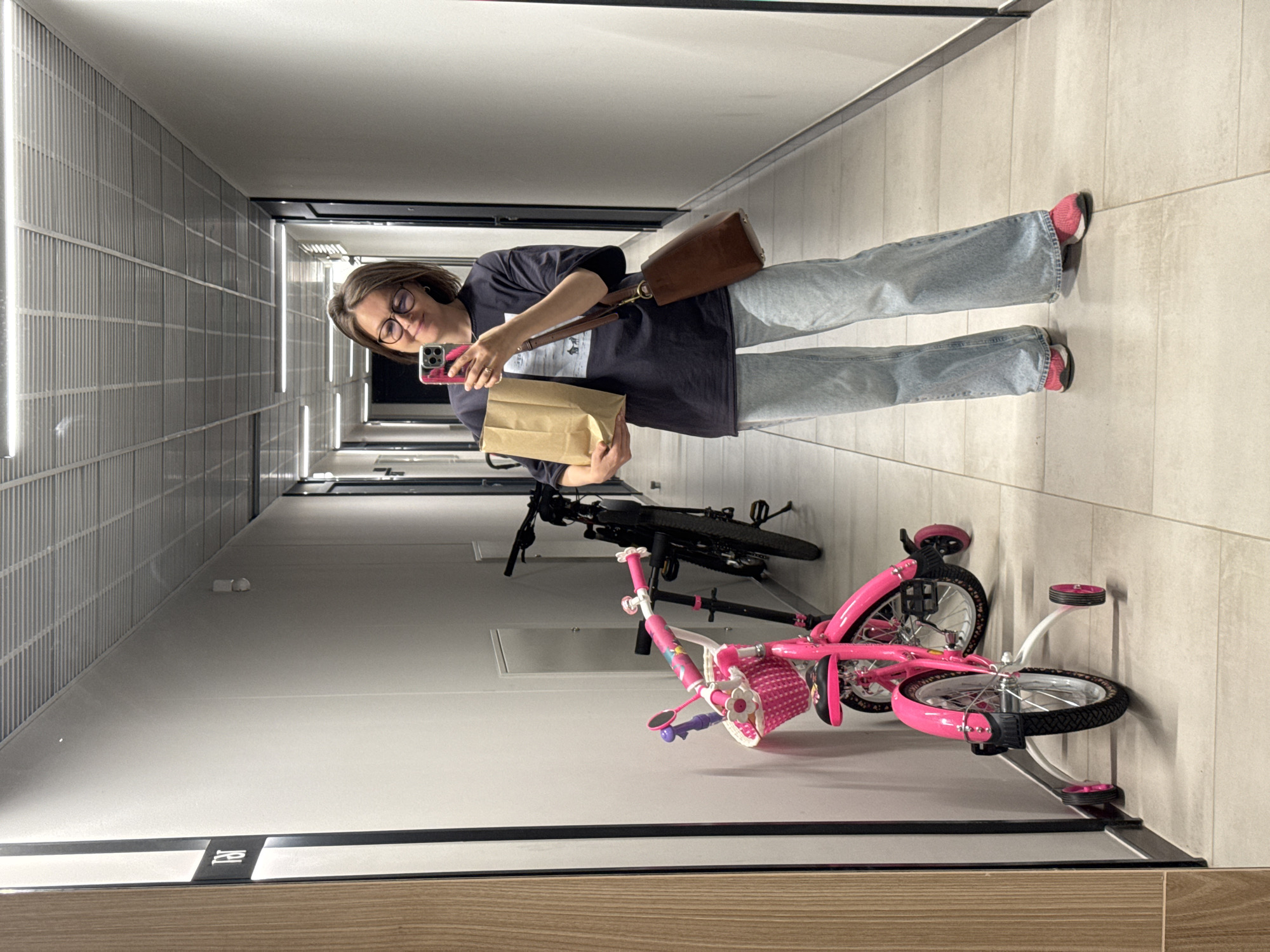The image shows a woman in a hallway, holding a paper bag and a smartphone. She is wearing a dark t-shirt, light blue jeans, and pink athletic shoes. A brown leather shoulder bag is slung across her body. To her left, there is a pink children's bicycle with training wheels, and behind it, a black bicycle. The hallway has light-colored walls and a tiled floor. A sign with the letters "FB" is visible on the left wall. The ceiling features fluorescent lighting and a grated metal structure. The woman is looking at her smartphone, which she is holding up to take a selfie in a mirror. Based on the visible sign and the typical architecture of apartment buildings in Russia, this scene is likely a residential building hallway in Izhevsk, Russia.