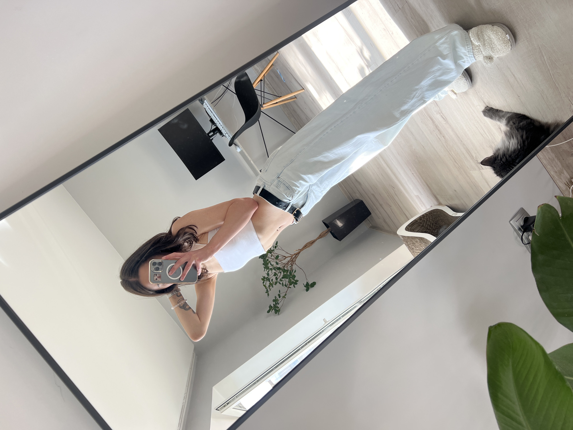 A young woman is taking a mirror selfie in what appears to be a modern, well-lit apartment. She is wearing a white crop top and light blue, wide-leg jeans with a black belt. Her hair is dark and falls to her shoulders. She is posing with one arm raised behind her head. In the reflection of the mirror, a desk with a computer monitor and keyboard is visible, along with a black ergonomic chair and a tall plant in a black pot. Sunlight streams in from a window, casting shadows on the wooden floor. A grey cat is lying on the floor near the woman's feet. In the foreground of the mirror's reflection, large green leaves of a houseplant are visible. The overall impression is one of a casual, everyday moment in a domestic setting. The image does not provide specific details to determine the location within Ternopil, Ukraine.