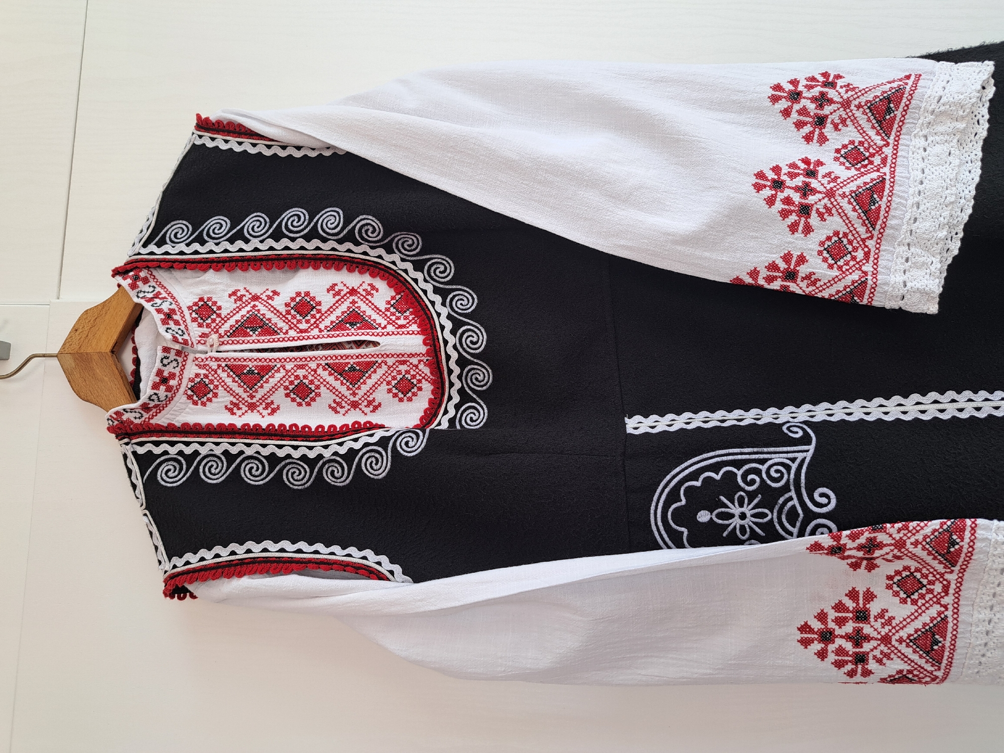 The image shows a traditional Bulgarian folk costume laid flat on a white surface, partially hanging from a wooden hanger. The costume features a black vest adorned with white embroidery in spiral and floral patterns. The sleeves and yoke are white linen with red and black geometric embroidery, finished with white lace trim. The scene is indoors under even lighting.