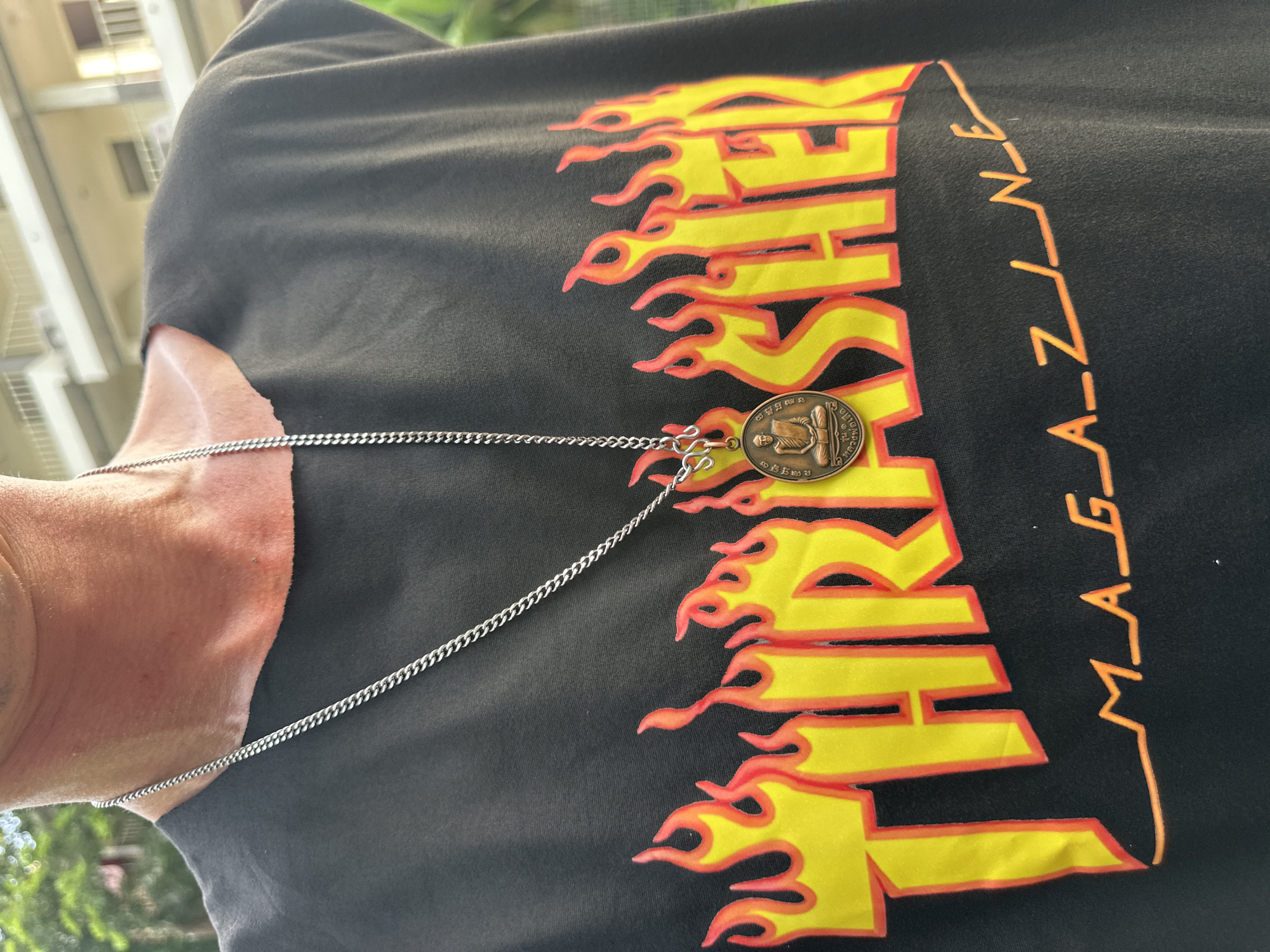 In Karon, Thailand, a person wears a black t-shirt with the "Thrasher Magazine" logo printed in yellow with red flames. A silver chain necklace with a small, oval-shaped medallion featuring a seated Buddha figure is worn over the shirt. The background shows a blurred residential building.