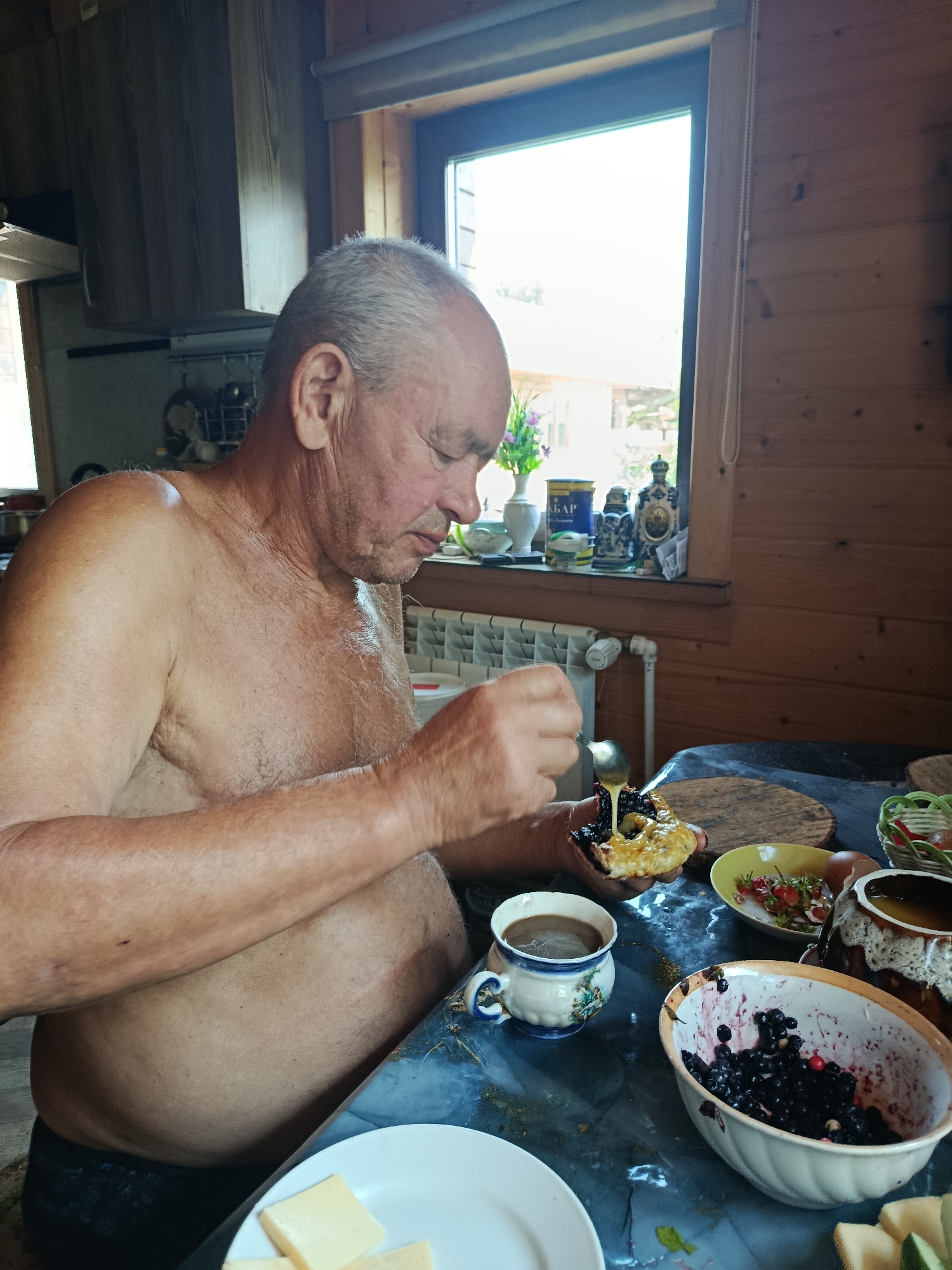 The image captures an indoor scene, likely a kitchen or dining area, within a wooden-paneled room that appears to be a dacha or rural dwelling in Vareya, Russia. A middle-aged to older man, shirtless and wearing dark underwear, is seated at a table. He is actively engaged in preparing a piece of food, possibly bread or a pastry, by spooning a dark, berry-like substance and a golden liquid, likely honey or syrup, onto it.
The table is covered with a dark blue tablecloth and displays various items, including a white and blue patterned teacup filled with coffee, a bowl overflowing with dark berries, a yellow bowl containing strawberries, a ceramic pot with a handle, and several slices of cheese on a white plate. In the background, a window offers a glimpse of the outdoors with greenery and possibly some buildings. A wooden cabinet is visible on the left, and the wall on the right is made of vertical wooden planks. A radiator is also present beneath the window. The overall atmosphere suggests a simple, informal meal.