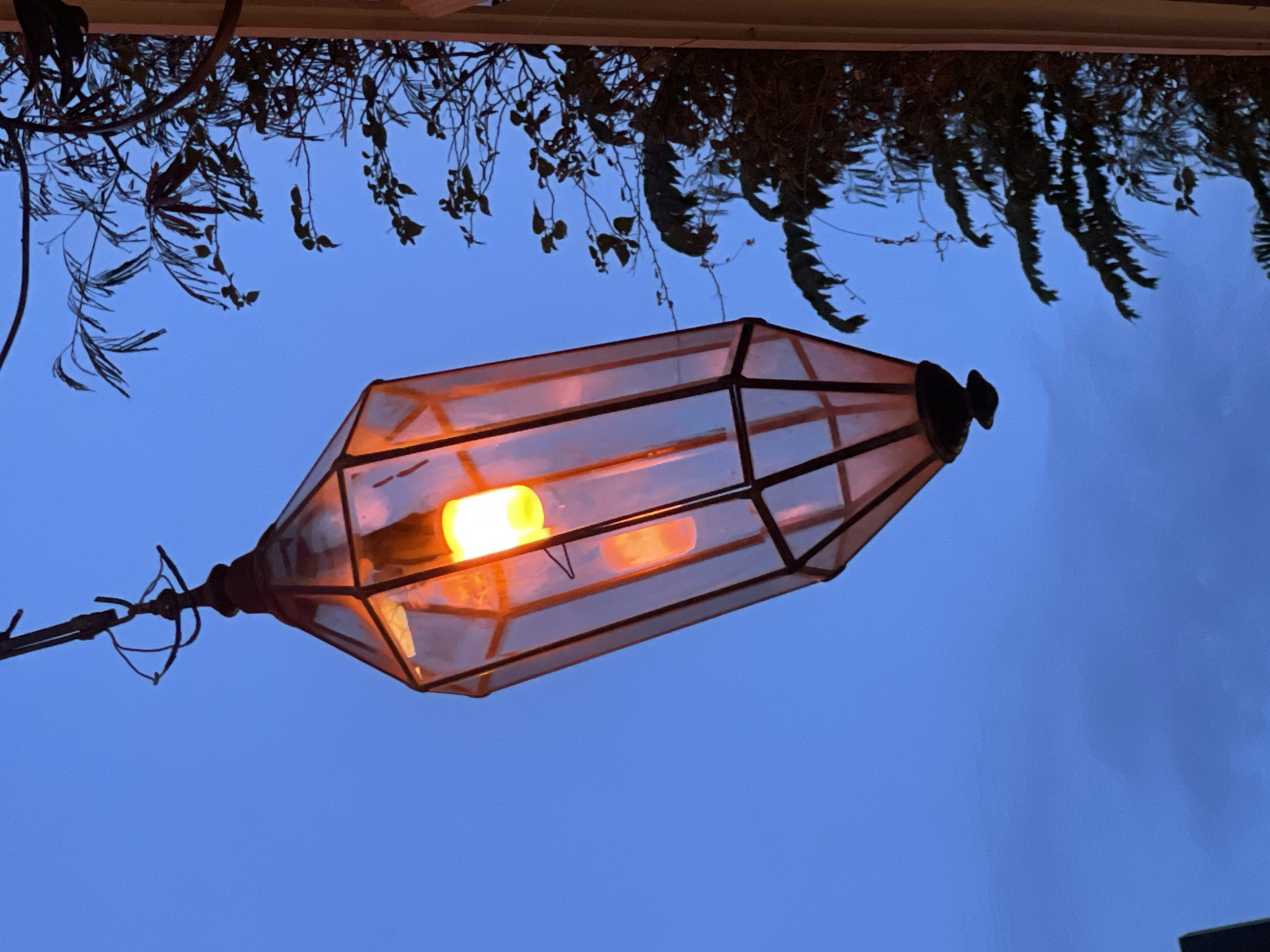 An illuminated lantern hangs from a dark metal fixture against a deep blue twilight sky. The lantern, with its hexagonal panels and dark metal frame, casts a warm, orange glow from an internal light bulb. The glass panels of the lantern appear to be tinted a reddish-orange. In the upper right portion of the frame, a dark silhouette of tree branches and leaves is visible against the sky. To the right of the lantern, a portion of a pale, vertical structure, possibly a wall or pillar, is present. The overall scene suggests an outdoor setting at dusk. The location is identified as Pattaya City, Thailand.