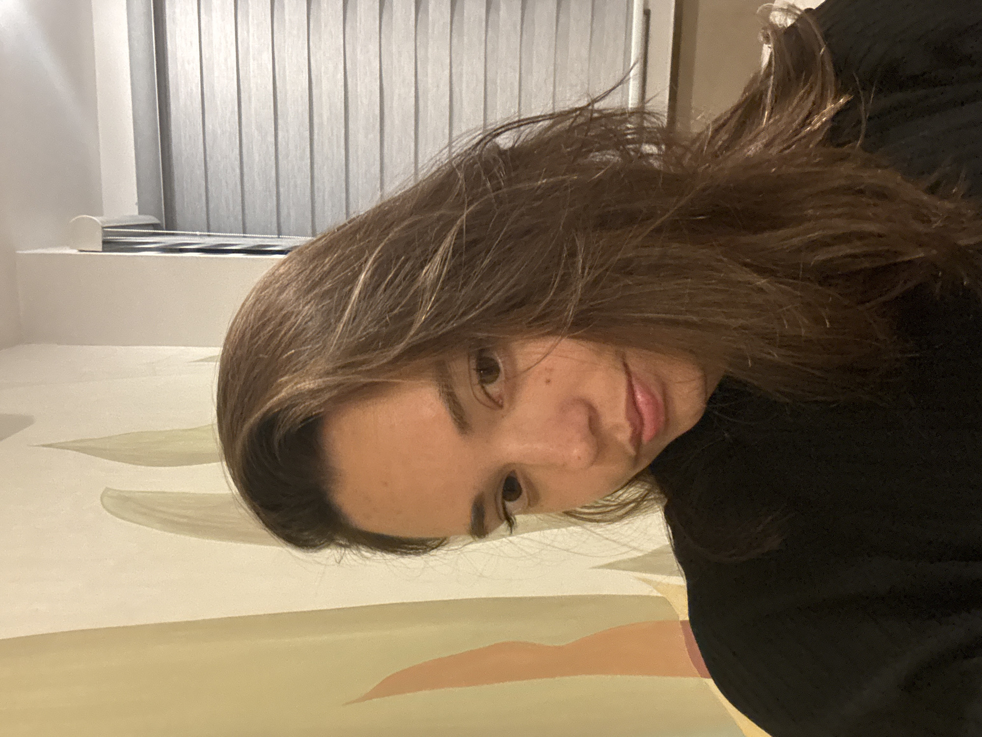 A person with brown hair is seen indoors with light filtering through window blinds in the background. The camera angle is tilted, showing the person's face close-up. The background consists of a light-colored wall. There is no available data to indicate this photo was taken in San Bernardino Tlaxcalancingo, Mexico.