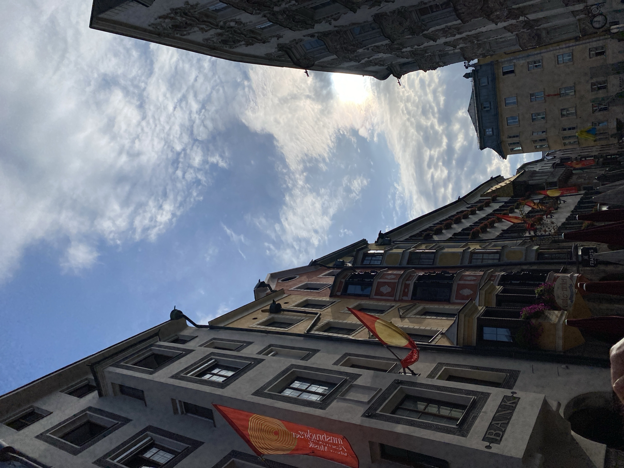 This image captures a low-angle view of the historic streets of Innsbruck, Austria, under a partially cloudy sky. The architecture is a key feature, with narrow, colorful buildings lining a street. The building on the left is predominantly gray, with a prominent "BANK" sign etched into its facade. Two flags hang from this building: a large red banner with a spiraling yellow design and text, and a smaller red and yellow striped flag. Further down the street, other buildings exhibit a variety of colors and architectural details, including bay windows adorned with flowers and ornate stonework on the building to the right. The sun is visible through the clouds, casting a gentle glow and a subtle rainbow effect. No people are clearly visible in the frame. The scene suggests a bustling European city with a rich history, likely within the Old Town of Innsbruck, known for its well-preserved medieval and Baroque architecture.