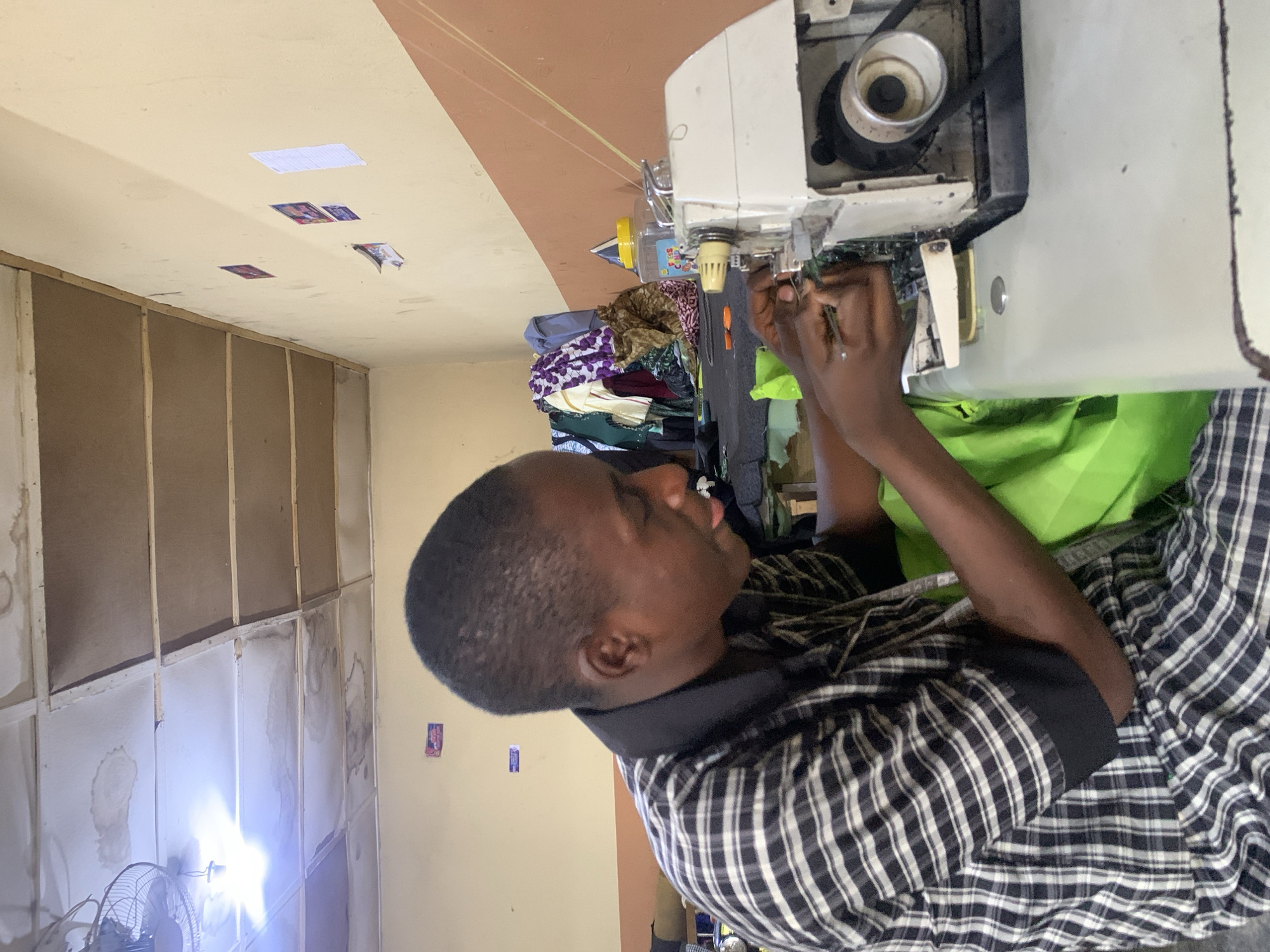 A man is seated at a sewing machine, seemingly repairing it. He is wearing a plaid shirt and has dark skin. The sewing machine is white and appears to be a heavy-duty industrial model. Various fabrics are piled nearby. The location appears to be a workshop or small tailor shop. The walls are unfinished, with evidence of water damage and attached posters. A fan is visible. This likely is an economical home business location. Although the location of Jos, Nigeria is requested, there is nothing visible to confirm location.
