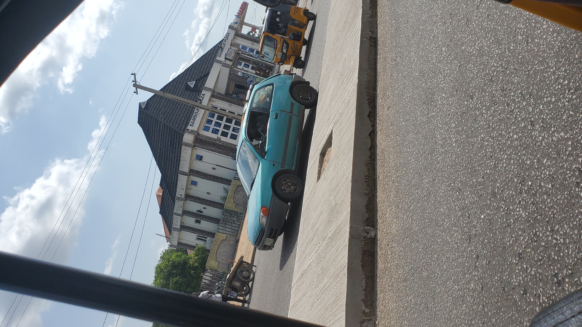 The image depicts a street scene in Jalingo, Nigeria. The main subjects are a light blue car, a yellow and black auto-rickshaw, and a multi-story building. The car, a sedan, is parked on the side of a paved road, angled slightly towards the viewer. The auto-rickshaw is behind the car, also parked. The building, labeled "SAMBOJO PLAZA" in blue lettering, is modern with a dark, angled roof and multiple windows. The sky is partly cloudy, suggesting daytime. The overall scene appears to be a typical urban street, possibly a commercial area given the building's signage. There is a concrete barrier or wall in the foreground on the right side of the image. The image is taken from a slightly elevated perspective, likely from a vehicle. The lighting suggests it is a sunny day.