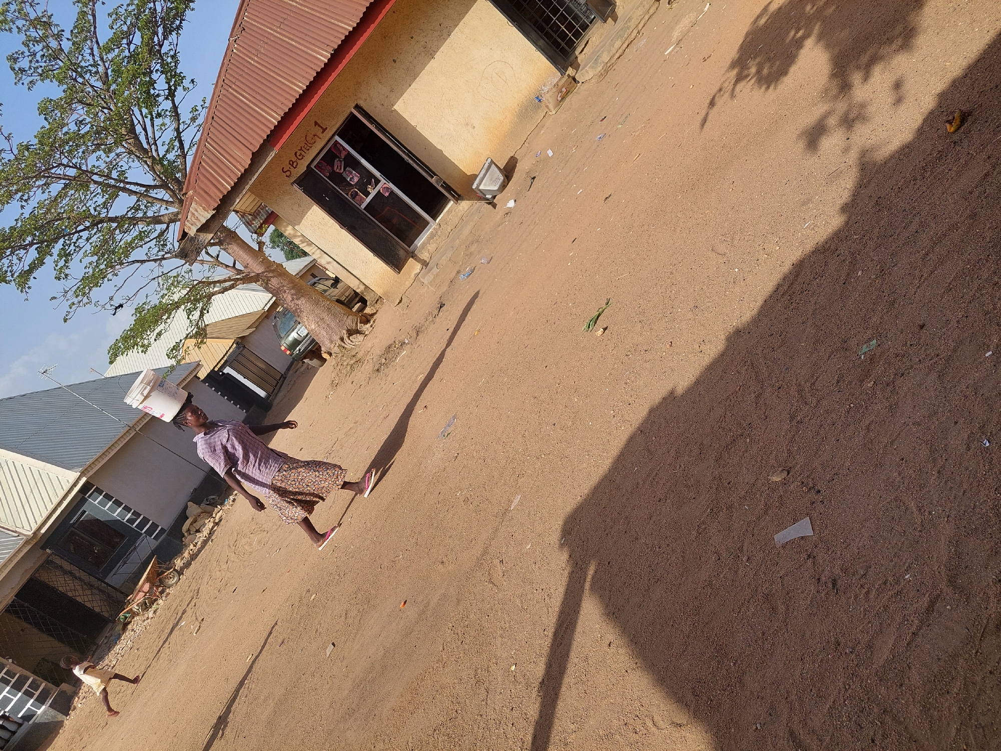 The scene depicts a dusty street in Barakin Angundi, Nigeria. A woman wearing a patterned dress and flip-flops walks while balancing a white bucket on her head. A young child in a light-colored dress walks in the same direction. To the left, there is a building with a corrugated metal roof. In front of the building, there's a large tree. Further down the street is another building. Litter is scattered on the ground. A large shadow from a tree dominates the right side of the frame.