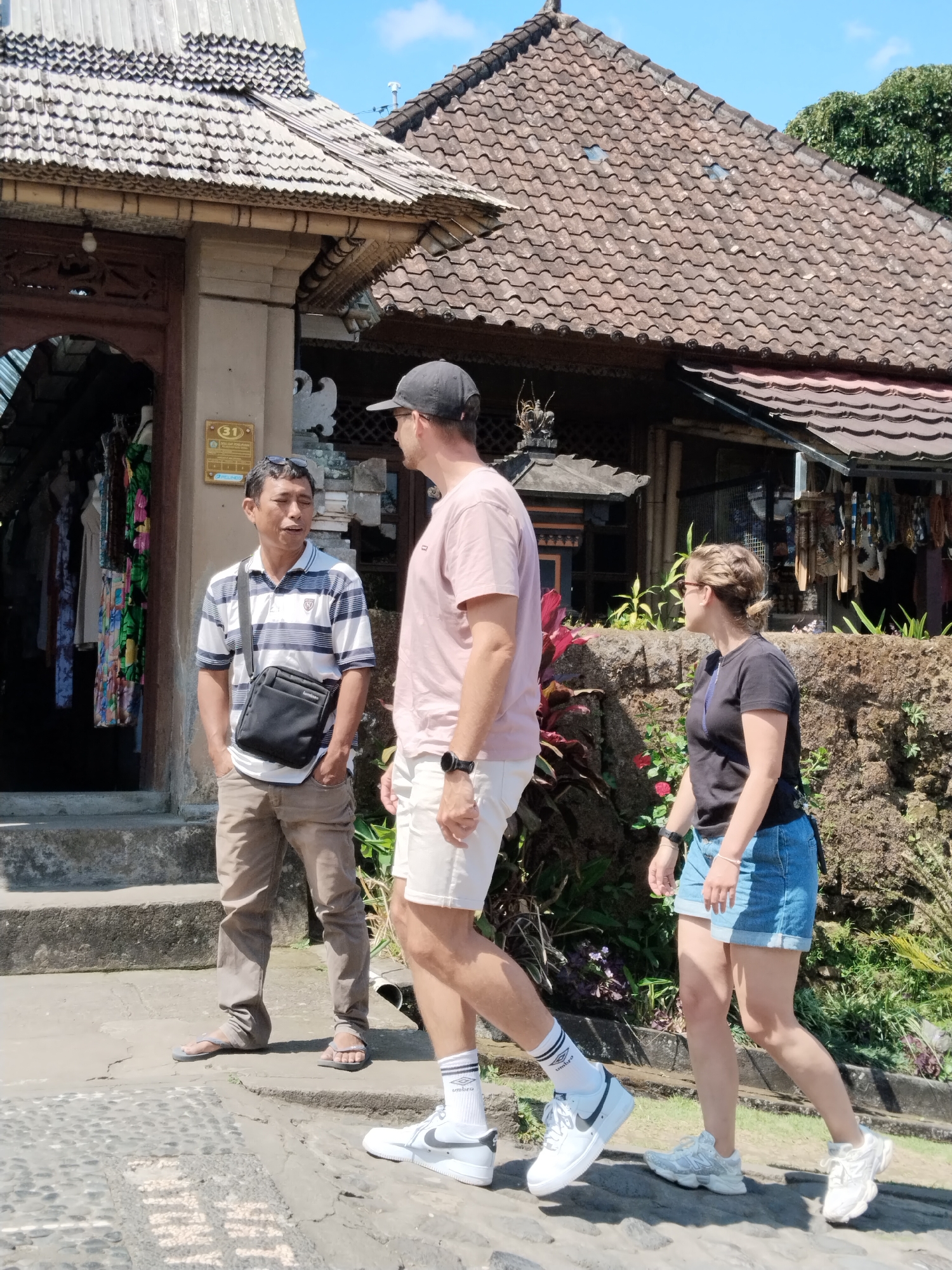 Three people are standing on a paved path in front of a building in Bangli, Indonesia. The person on the left is a man wearing a striped polo shirt, khaki pants, and sandals. He has a black crossbody bag. He is speaking to a man in the center of the image, who is wearing a pink t-shirt, khaki shorts, white socks, and white sneakers. The man in the center is wearing a black baseball cap and a watch on his left wrist. To the right of these two men is a woman wearing a black t-shirt, denim shorts, and white sneakers. She is looking to her right. The building behind them has a thatched roof and ornate wooden details. There are shops visible inside the building, displaying colorful clothing. A stone wall with plants is to the right of the building. The ground is paved with stones and concrete. The sky is blue with some clouds.