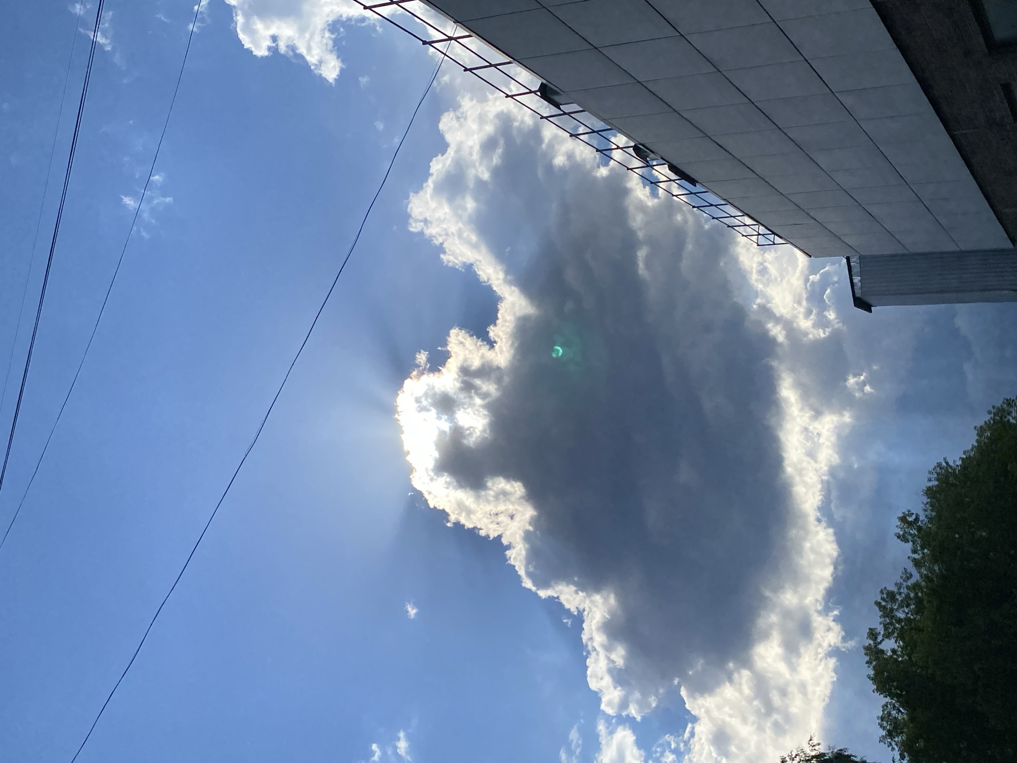 This media file captures a view of the sky from Votkinsk, Russia. The scene features a dramatic sky with a large, dark cloud dominating the center. Sunlight pierces through the edges of this cloud, creating visible sunbeams. Within the cloud, there is a bright, greenish-blue lens flare, suggesting the presence of the sun. A building with a modern, tiled facade and dark, horizontal window frames extends into the upper right of the frame. A few thinner, wispy clouds are scattered in the blue sky. In the lower left, power lines are visible, stretching diagonally across the frame. In the lower right, the tops of green trees can be seen. The lighting indicates it is likely daytime, with the sun partially obscured by the clouds, creating a mix of bright and shadowed areas. The overall impression is of a dynamic and somewhat dramatic sky. No people are visible. There is no visible text.