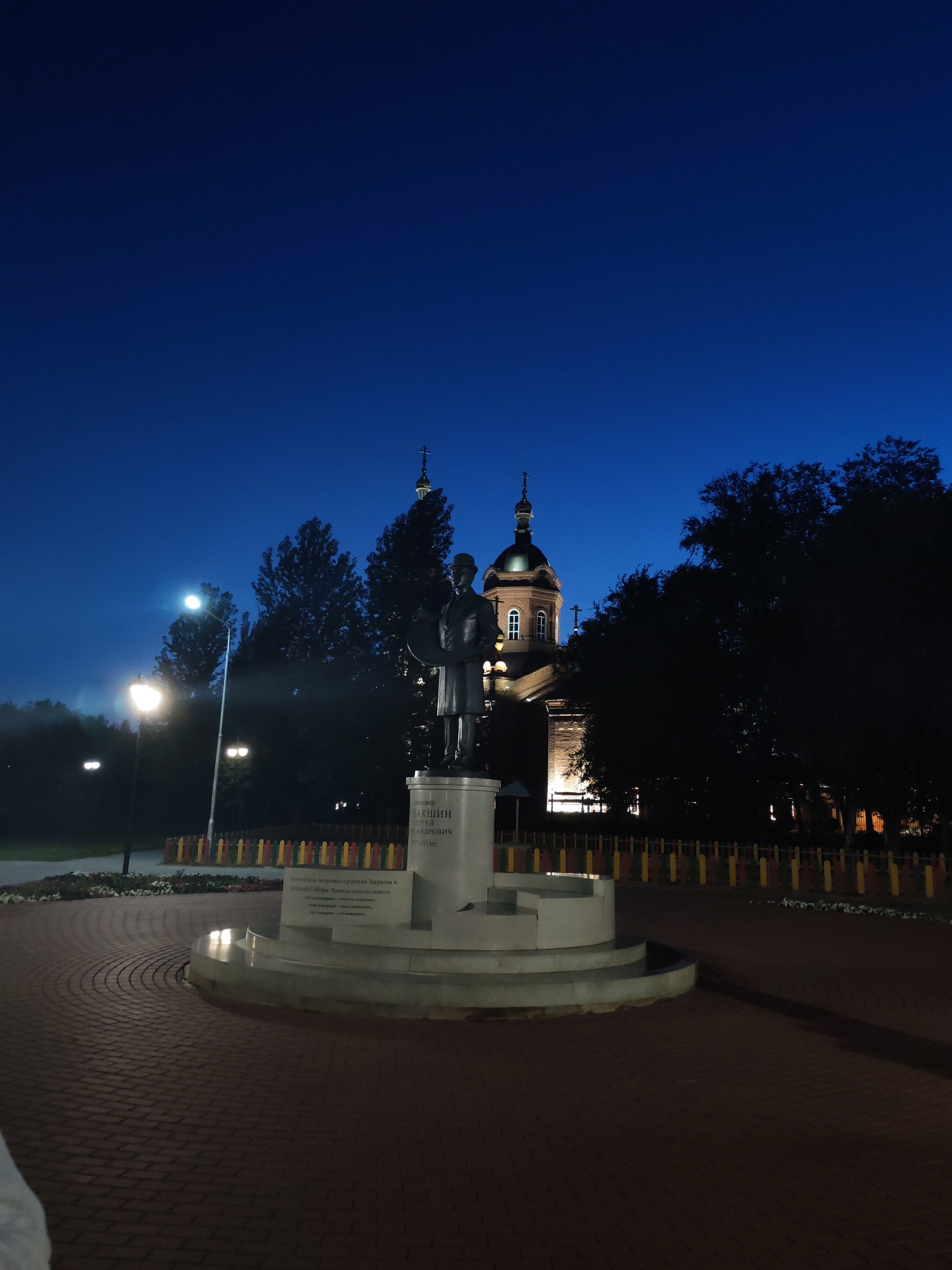 The image shows a statue of a man in front of a church at night. The statue is made of bronze and depicts a man in a suit holding a tablet. The church is made of brick and has a dome. The sky is dark blue, and there are trees in the background. The ground is paved with bricks. The statue is lit by spotlights, and the church is also illuminated. There is a fence in front of the church, and some flowers in front of the fence. The image is taken from a low angle.