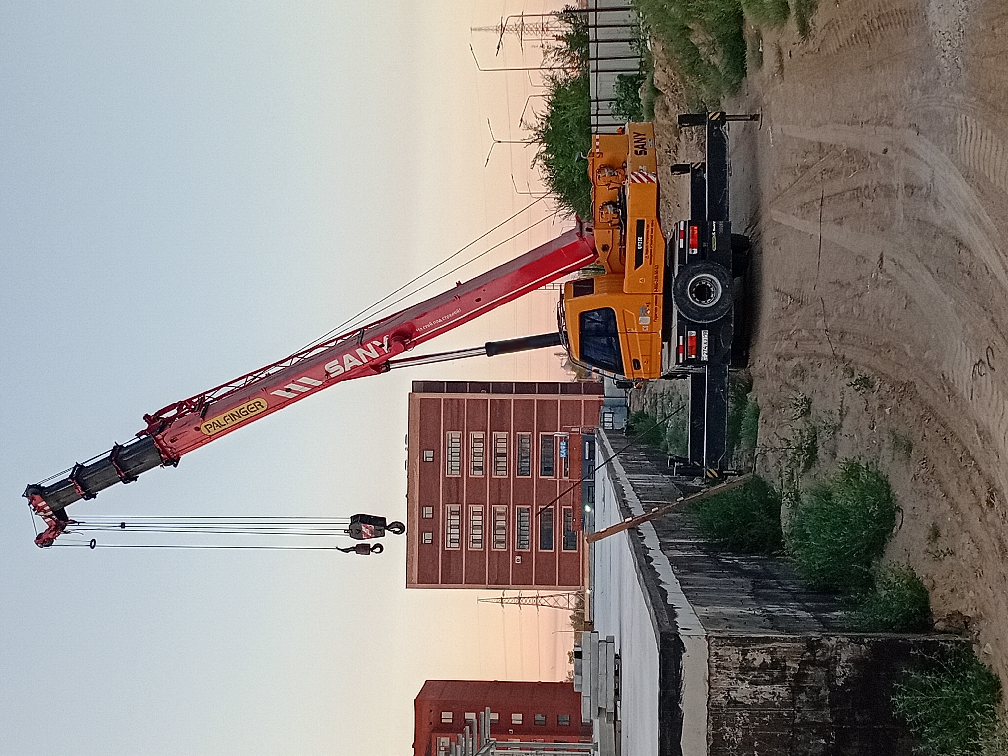 An orange and red SANY crane is parked on a dirt road at a construction site. The crane's boom is extended and raised, with a hook assembly suspended in the air. In the background, there are two brick apartment buildings, power lines, street lights, and a metal fence. The sky is clear with a pale orange hue, suggesting it is either sunrise or sunset. The area around the crane appears to be under construction, with piles of concrete slabs visible on a raised platform. The ground is covered in dirt and sand, with some overgrown vegetation. The image appears to have been taken during the day. The location is identified as Semey, Kazakhstan. There are no people visible in the image.