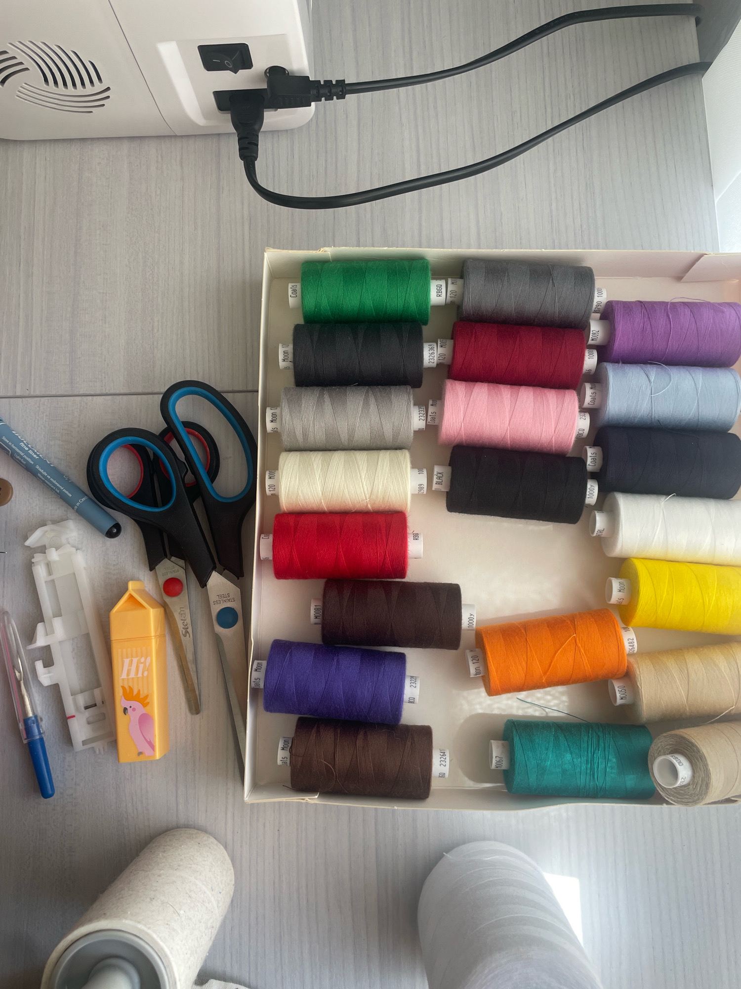 The image depicts a sewing machine with its power cord plugged in, placed on a grey wooden surface. Next to the sewing machine, there is a white box filled with spools of thread in various colors, including green, grey, purple, black, pink, red, orange, blue, and brown. Scattered around the box are sewing accessories such as scissors, a marker, a lint roller, and a bobbin winder. The scene appears to be a domestic setting, possibly a sewing room or a crafting area. The lighting suggests it is daytime, and there are no people visible in the image. The image does not provide any information about the city of Atarfe, Spain.