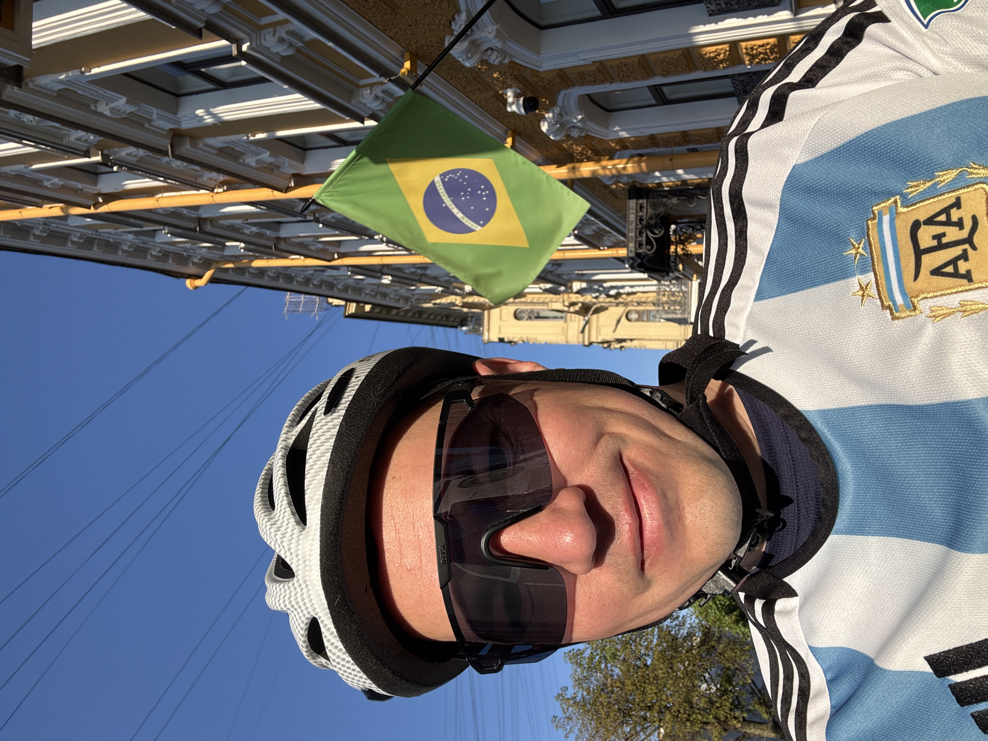 The image captures a bright, sunny outdoor scene, likely during daytime, with a person in the foreground and ornate architecture in the background. The main subject is a person, appearing to be male, wearing a white and light blue striped jersey, identifiable as the Argentina national football team kit, complete with the AFA (Argentine Football Association) logo and two gold stars. They are also equipped with a white and black cycling helmet and large, dark, wrap-around sunglasses, with the word "TRANI" subtly visible on the frame. The person has a neutral to slightly smiling expression.
In the background, a building with ornate classical architecture is visible, featuring beige or light yellow facades with intricate white decorative elements such as cornices and window frames. A small Brazilian flag, displaying its iconic green, yellow, and blue design with "ORDEM E PROGRESSO" (Order and Progress) written on its white band, hangs from this building. The sky is clear and blue, with some power lines and faint tree foliage also discernible. The person's attire suggests they are engaged in or prepared for cycling. The architectural style points to a European city, consistent with the provided location context of Saint Petersburg, Russia.
