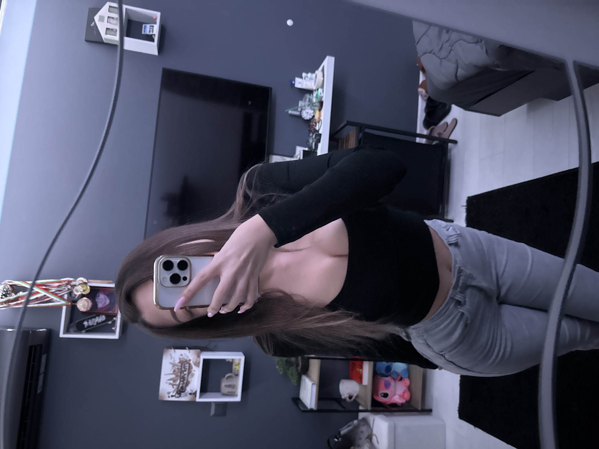 A person is taking a selfie in a mirror. The person has long brown hair, is wearing a black top and light gray jeans. The background of the image includes a TV, shelves with various items, a bed, and a black rug. The location appears to be indoors in a residential setting, but there's no information to confirm it's in Sofia, Bulgaria.