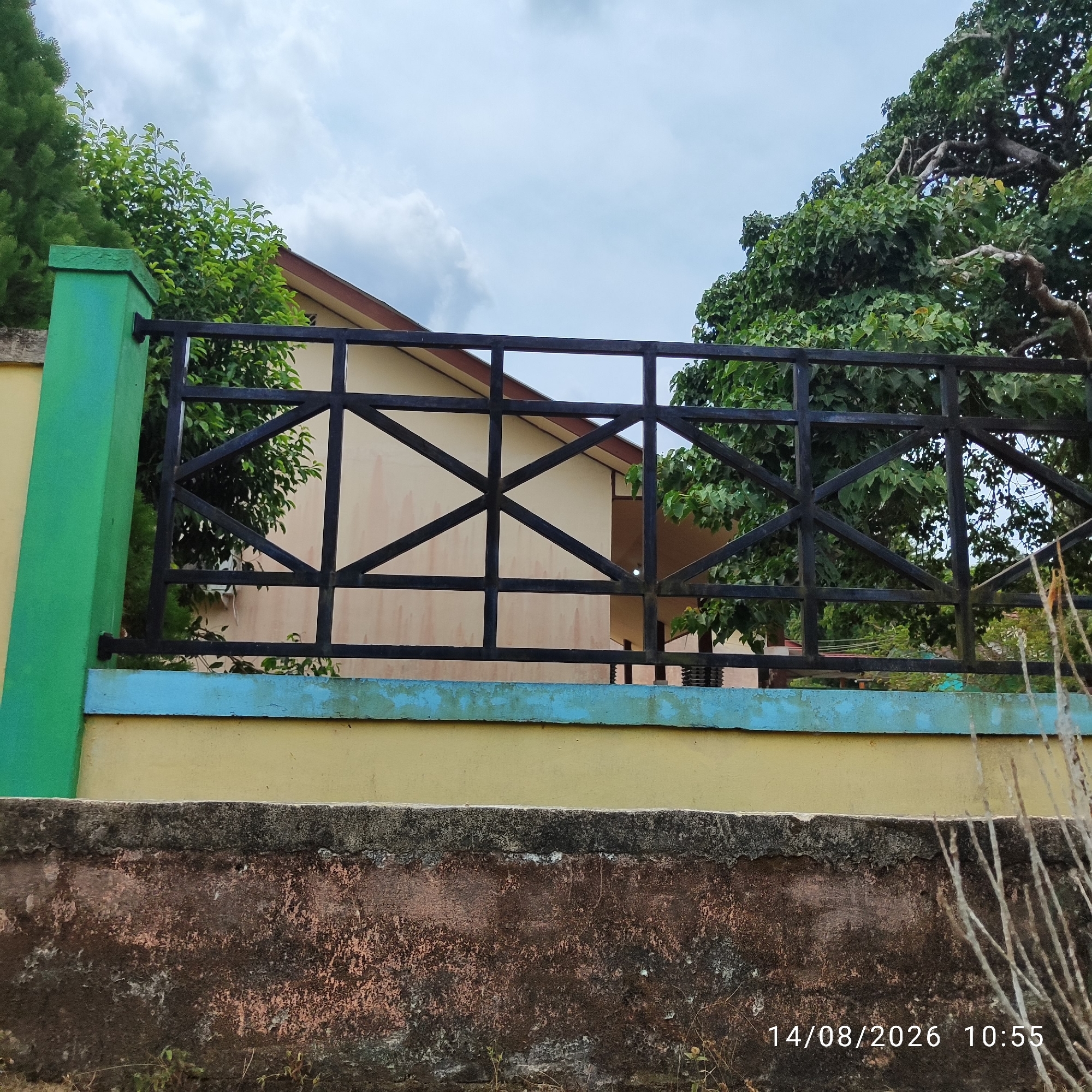 An outdoor, medium-shot, eye-level, full-color photograph shows a black metal fence in front of a light-colored building with a brown-trimmed roof. The fence is composed of horizontal bars and diagonal cross-braces. A bright green pillar is visible on the left side of the fence, and a yellow wall extends behind the fence. A thin blue strip runs along the top of the yellow wall, separating it from the fence. In the background, a dense canopy of green trees obscures parts of the building and the sky. The sky itself is overcast with light gray clouds. A weathered, dark brown concrete wall runs along the bottom foreground of the image, with sparse, dry vegetation growing in front of it. The date "14/08/2026" and time "10:55" are superimposed in white text in the bottom right corner.