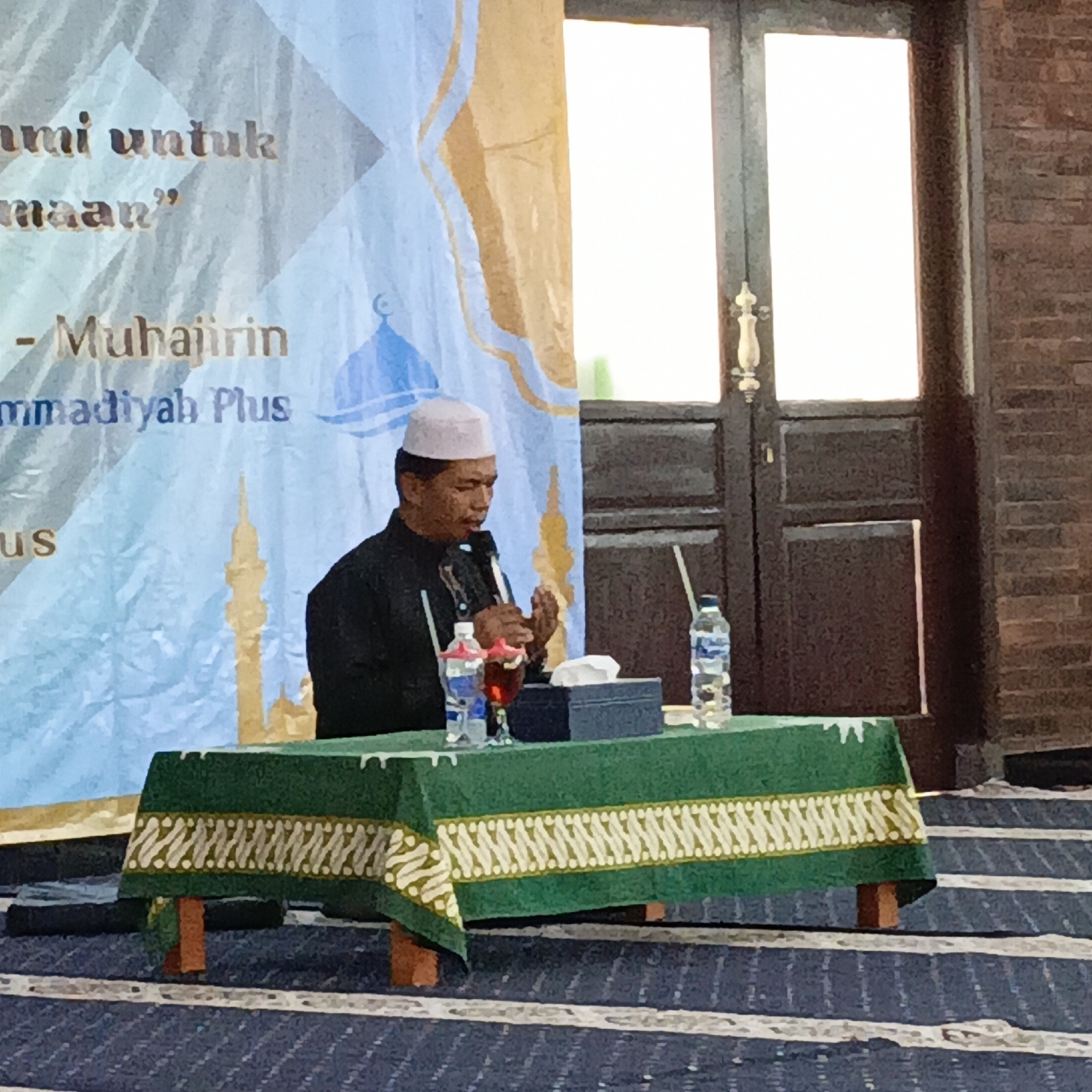 The image shows a man wearing a white peci (a cap) and a dark robe speaking into a microphone. He is seated behind a green tablecloth with a decorative yellow and white pattern, on which are placed a tissue box, a glass of red liquid, two bottles of water, and a microphone. The man appears to be in a room with a blue and gold banner in the background that features images of minarets and Islamic script. To the right of the banner are a pair of closed dark wooden doors with ornate handles, and a brick wall. The floor is covered with a blue carpet that has white lines running across it. The scene appears to be an event or gathering taking place in an indoor location. Based on the specific Islamic cultural context implied by the banner and attire, and the general architectural style, this scene could be taking place in a mosque, community center, or a formal event hall in a city like Salatiga, Indonesia, where such gatherings are common.