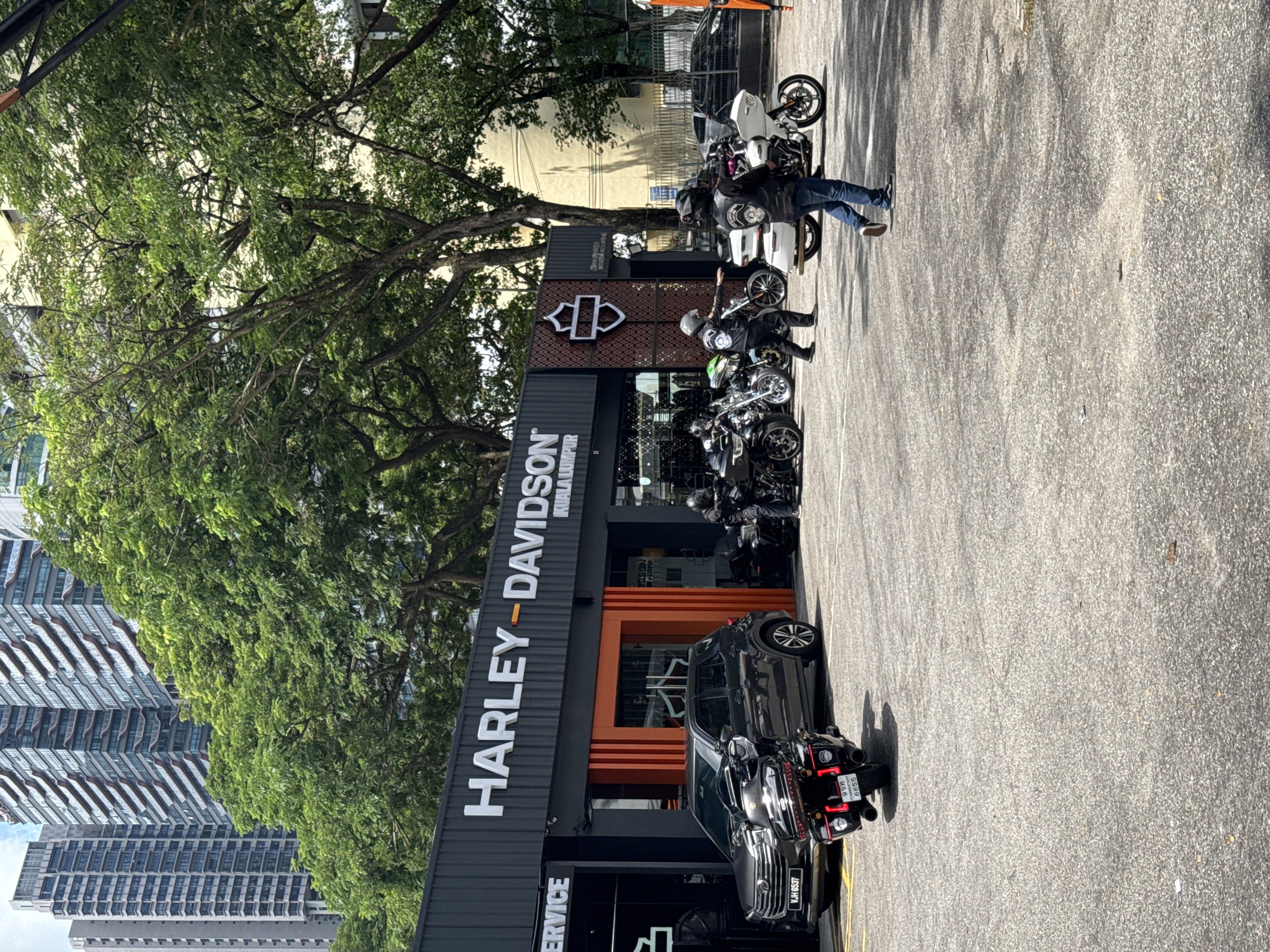 The image depicts a Harley-Davidson dealership in Kuala Lumpur, Malaysia. The dealership has a gray facade with orange accents and displays the Harley-Davidson logo prominently. Several motorcycles are parked in front of the store, along with at least one black car and another dark vehicle that partially obscures a bike. Several men, wearing motorcycle helmets and dark clothing, stand near the bikes. Trees and a modern skyscraper are visible in the background. The scene is outdoors on a sunny day.