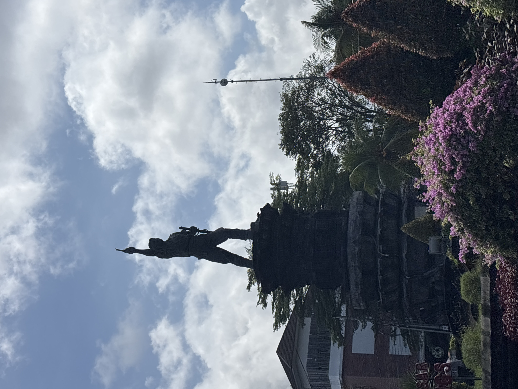 The image depicts a monument featuring a male figure pointing forward. The monument is positioned in a park-like setting with trees and flowering plants surrounding its base. Behind the monument is a multi-story building with a red-brick exterior. Above the monument is a partly cloudy sky. Based on the monument, the image is likely taken in Denpasar, Indonesia.