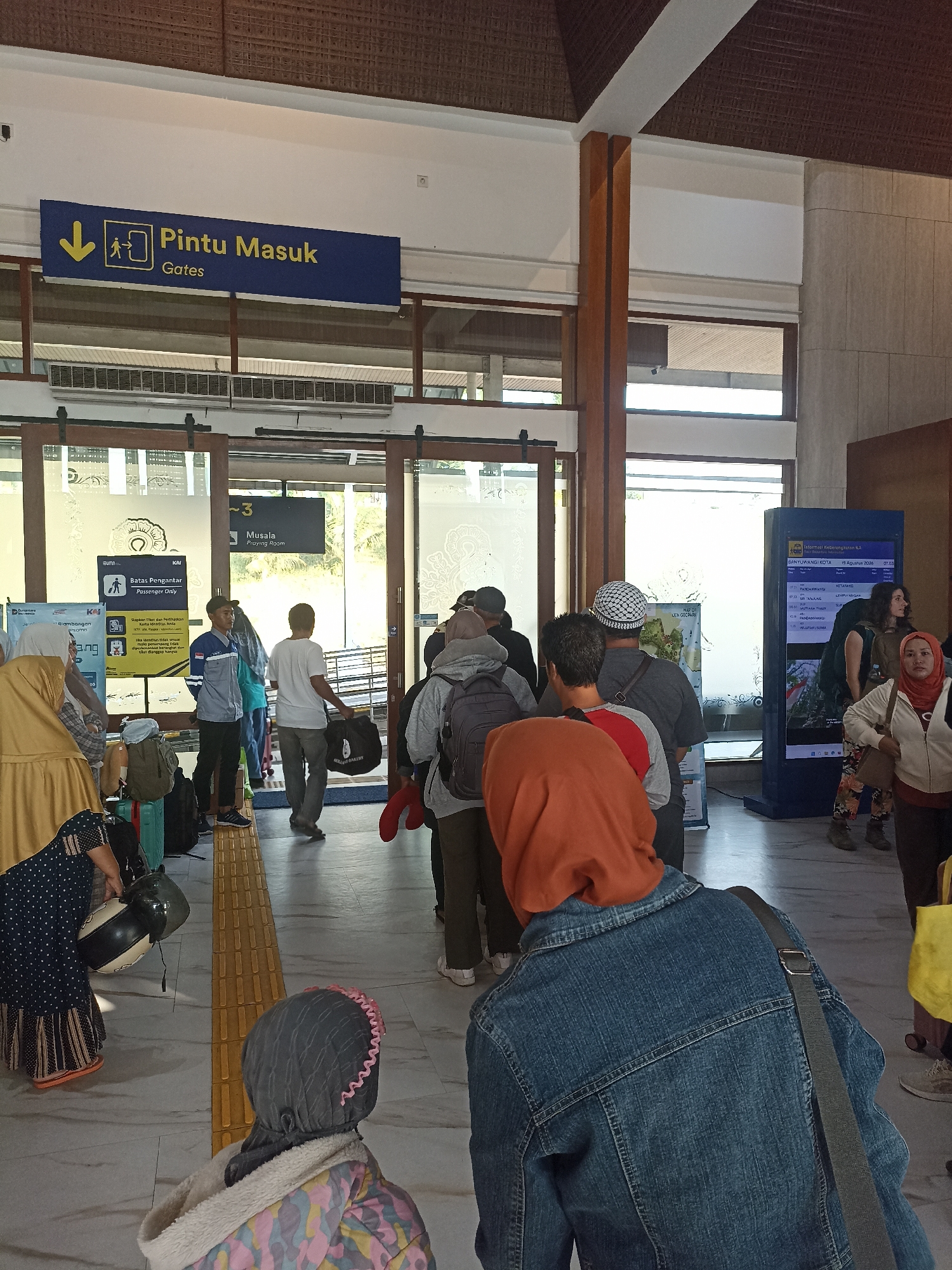 The image depicts the interior of a train station in Mojoagung, Indonesia. A group of people are queuing near a gate marked with "Pintu Masuk" and "Gates" in both Indonesian and English. The gate appears to be an entrance or checkpoint. Several individuals are carrying luggage, including bags and a helmet. The environment features a tiled floor, wooden pillars, and a ceiling with woven materials. A digital display board showing train information is visible on the right side of the frame, displaying "BANYUWANGI KOTA" and departure times. The overall scene suggests a busy passenger area within a transportation hub.